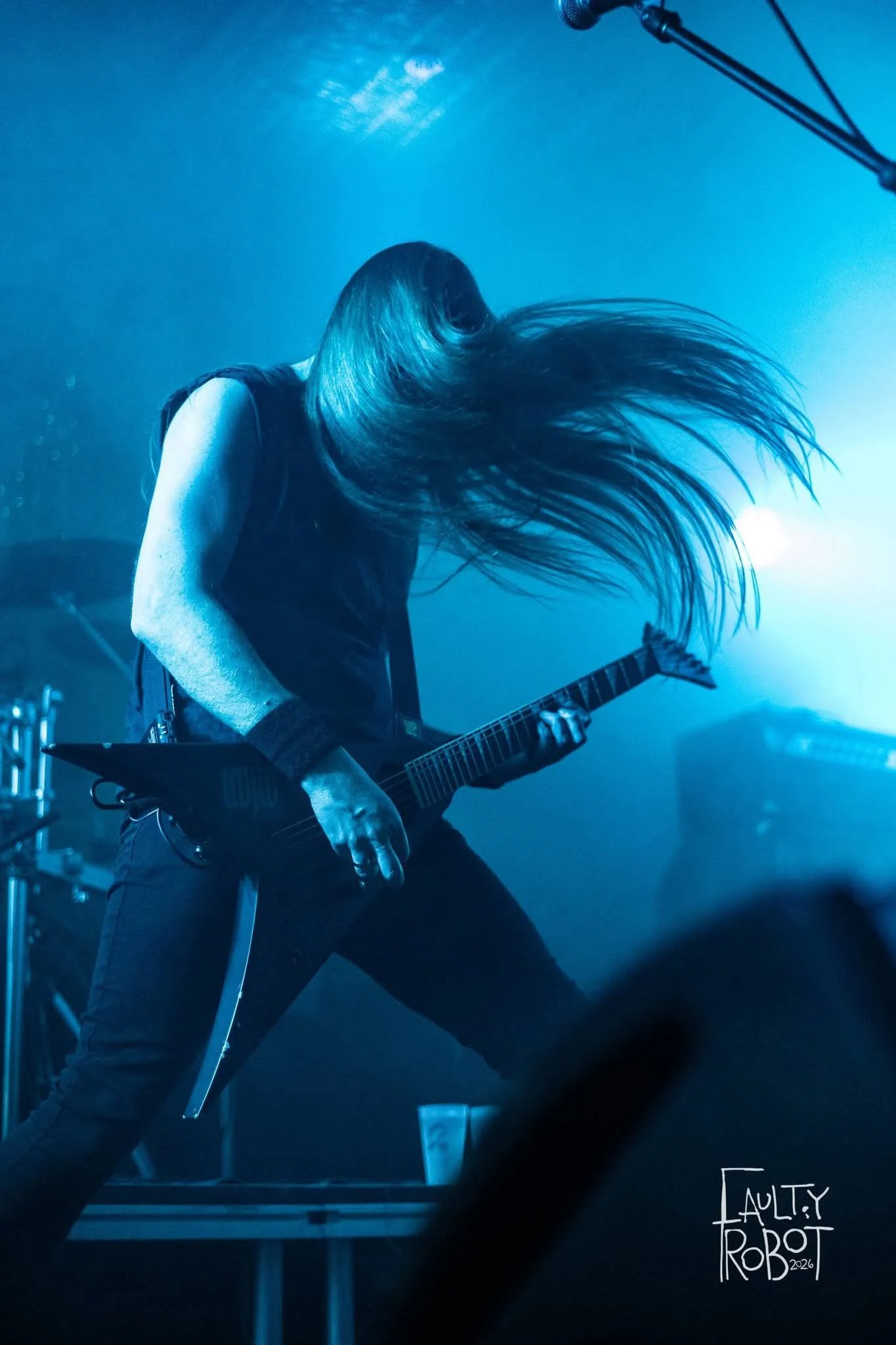 A musician with long hair playing an electric guitar on stage with blue lighting.