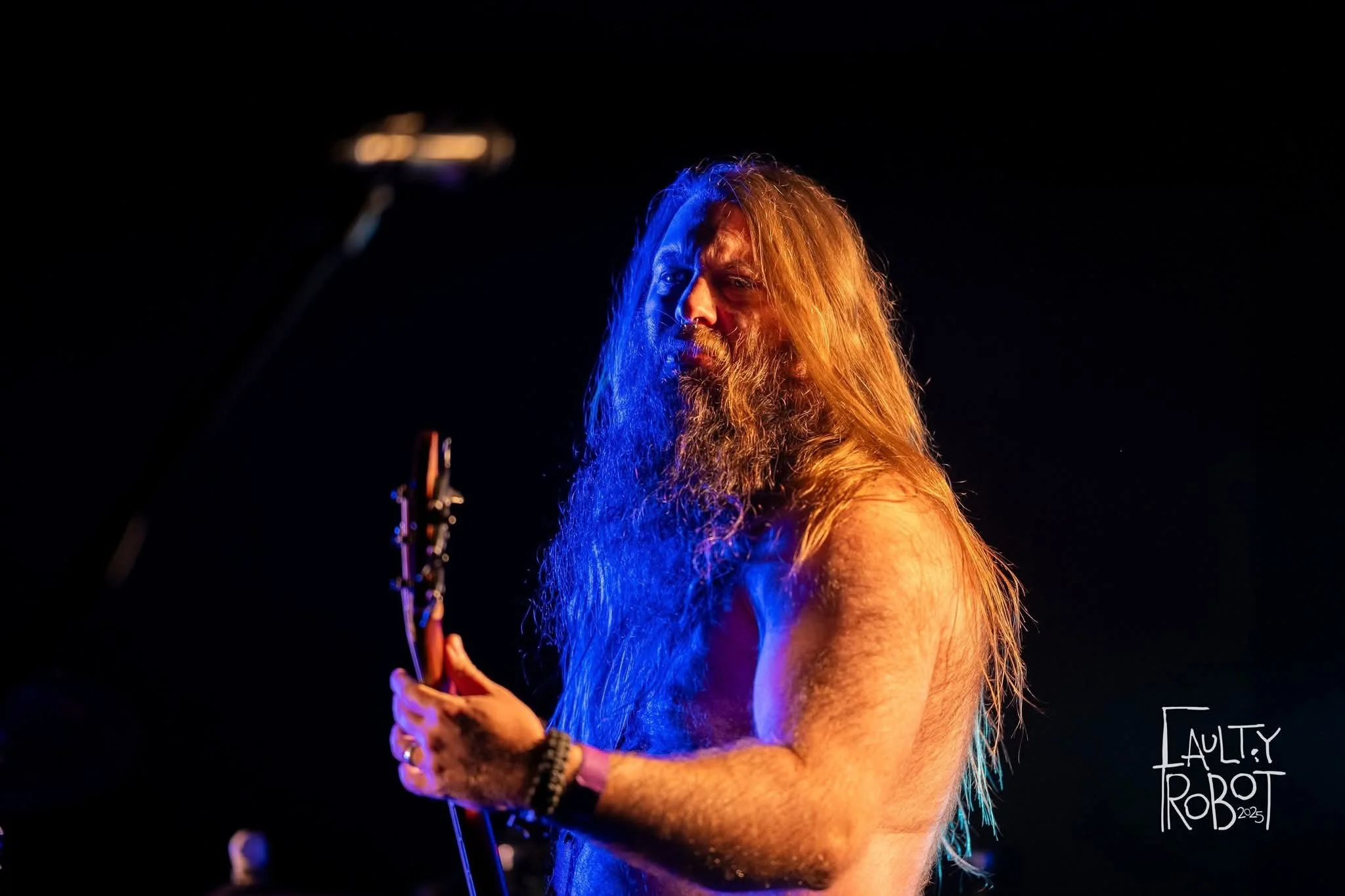 A shirtless long-haired man playing an electric guitar on stage, with dramatic lighting and a black background.