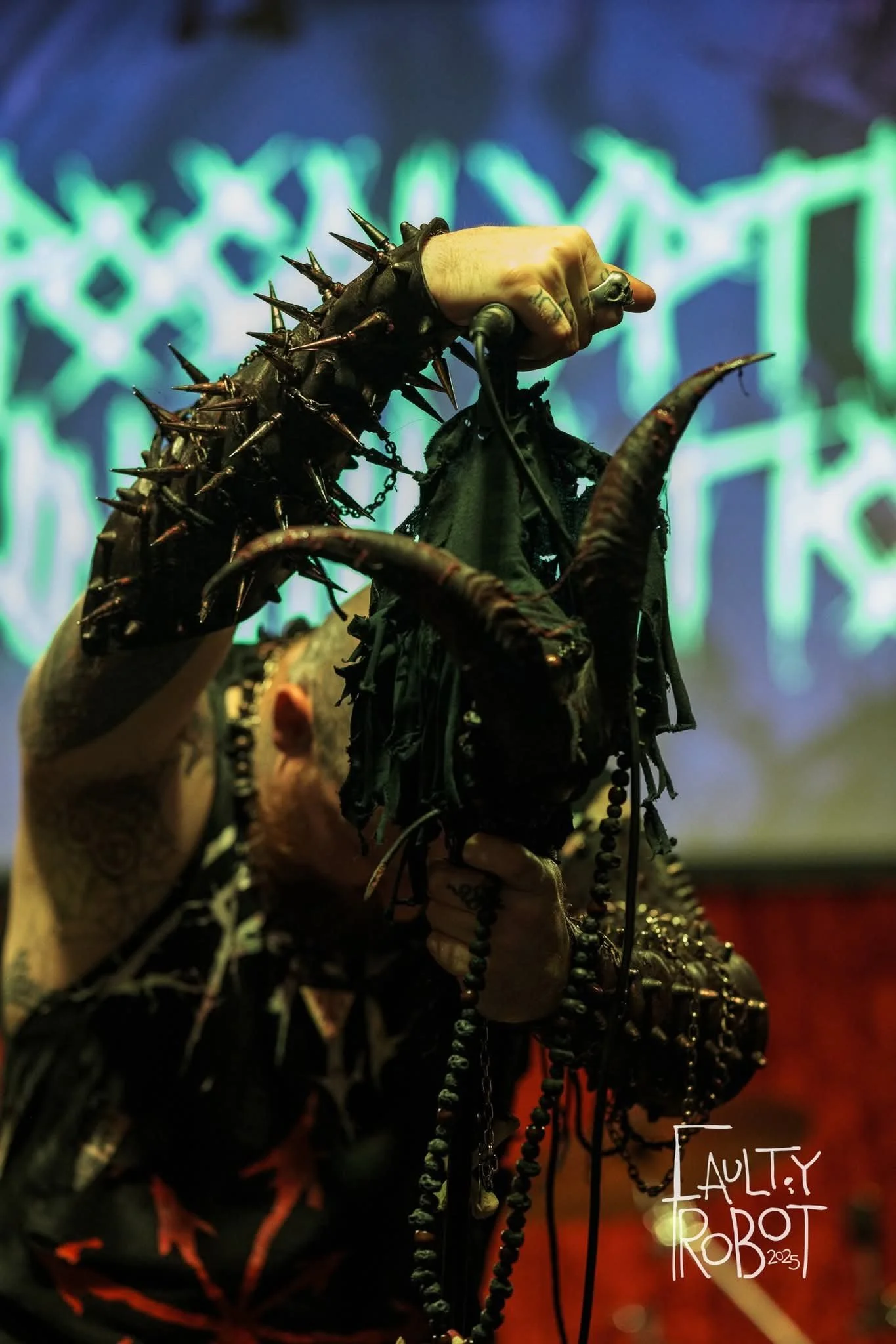 A performer holding a microphone in one hand and a necklace in the other, wearing black clothing with fringe and jewelry, with spiked accessories on their arm, under colorful stage lighting with a digital background.