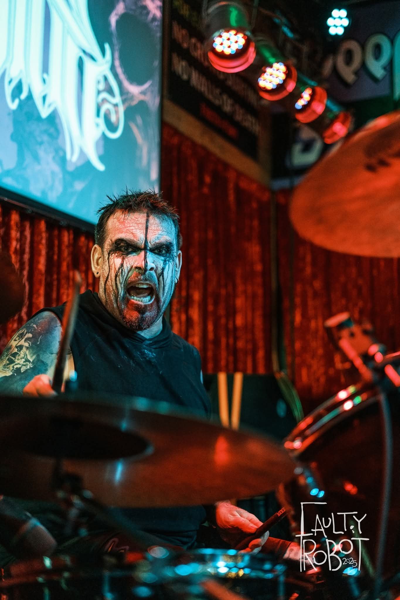 A drummer with face paint, looking angry or intense, performing on stage with colorful lighting and a red curtain background.