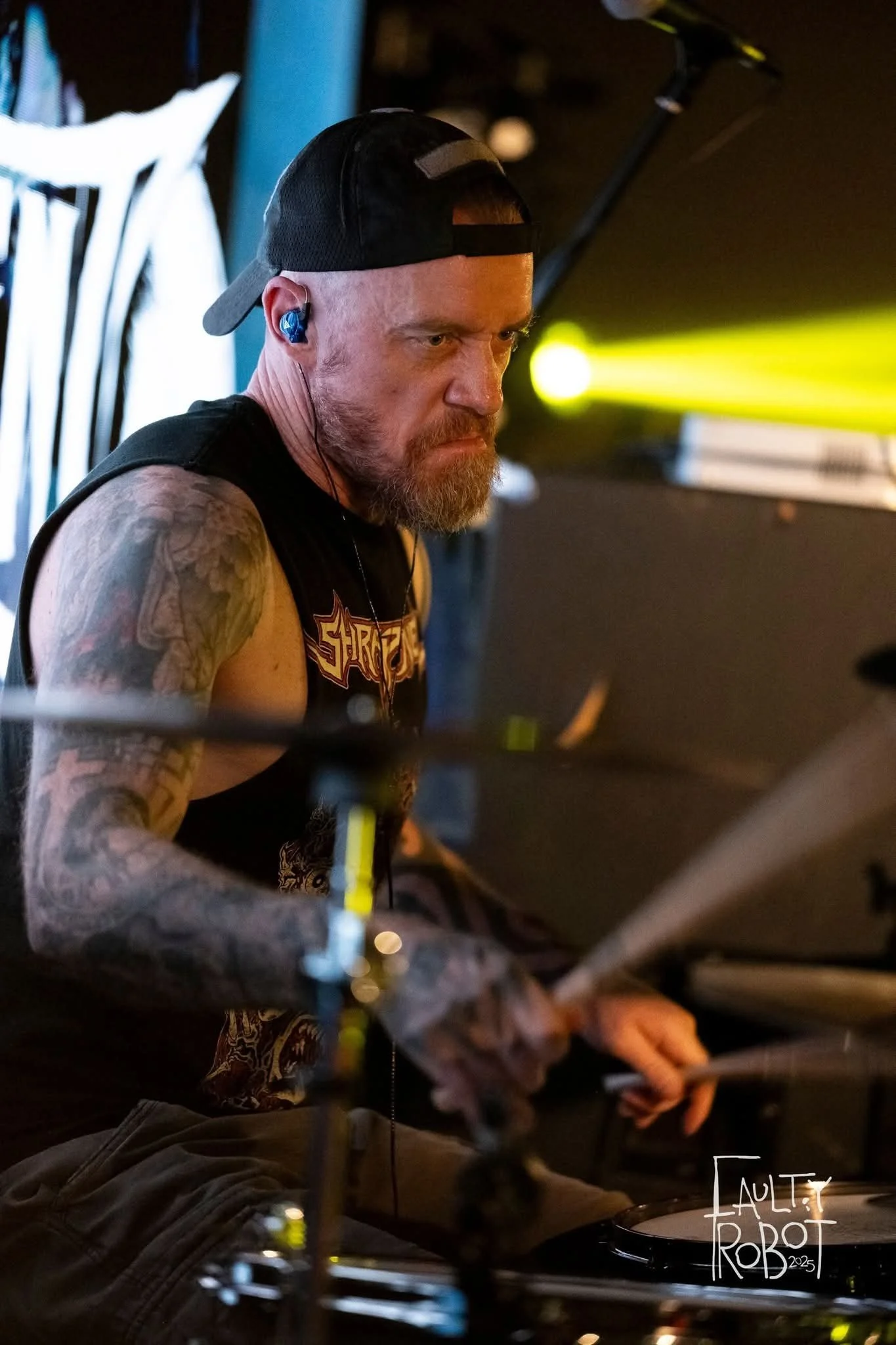 A male drummer with a beard, wearing a black baseball cap backwards, black sleeveless shirt, and wireless in-ear monitors, playing drums during a live performance with stage lights in the background.