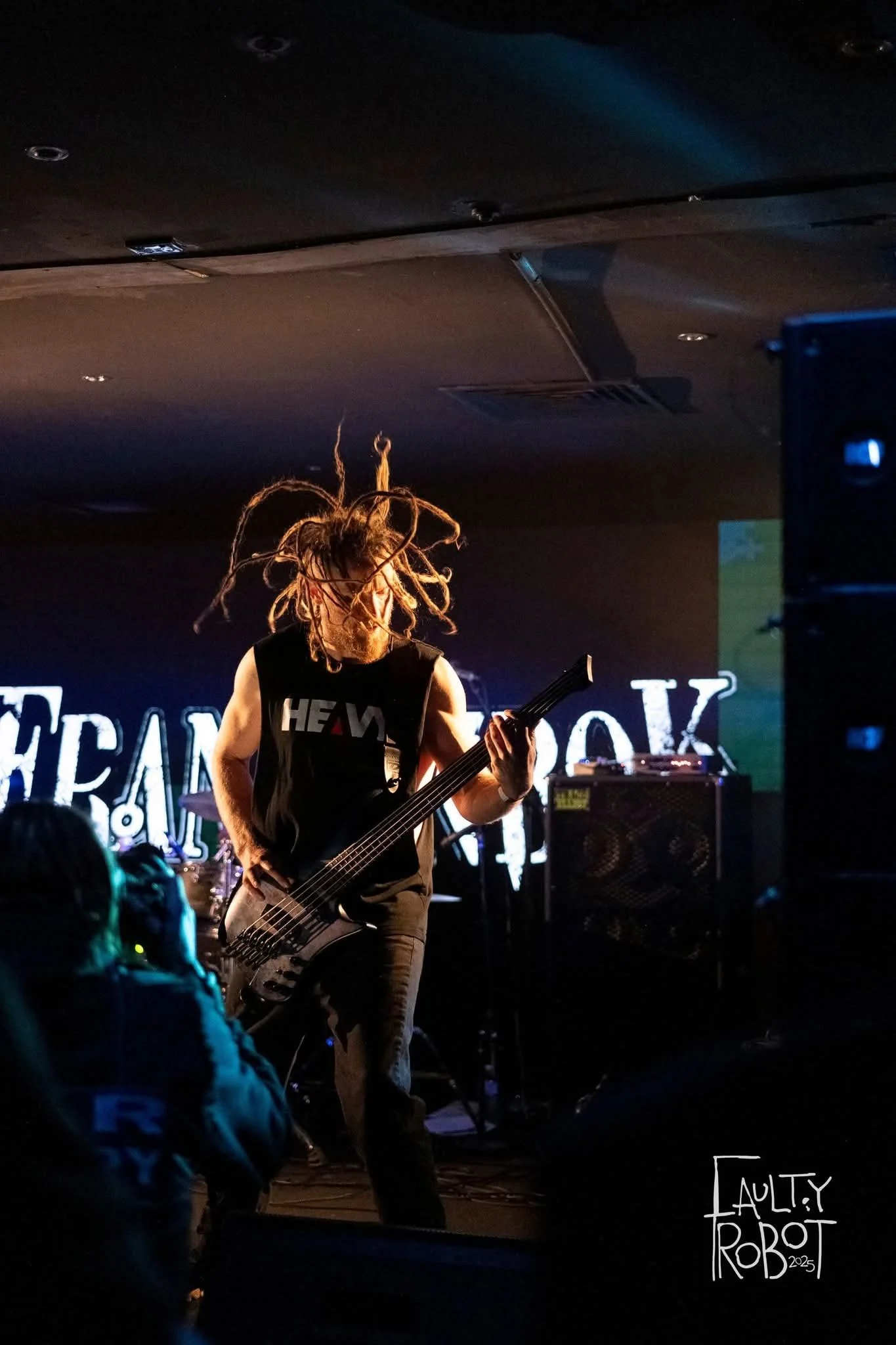 A musician with dreadlocks playing an electric bass guitar on stage during a live concert, with a dark background and stage lights, and an audience in the foreground.