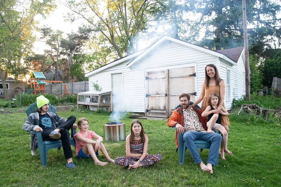 Family gathered outdoors in a backyard around a small fire pit, sitting on chairs and on the grass, with a white shed and trees in the background.