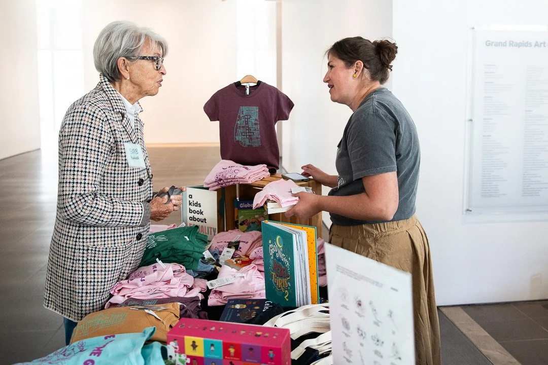 Two women talking at a merchandise table with books and T-shirts, indoors at an art gallery or museum.