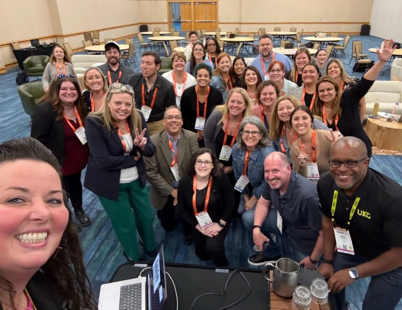Group of people at a conference or event, smiling and posing for a selfie in a spacious conference room with tables, chairs, and a carpeted floor.