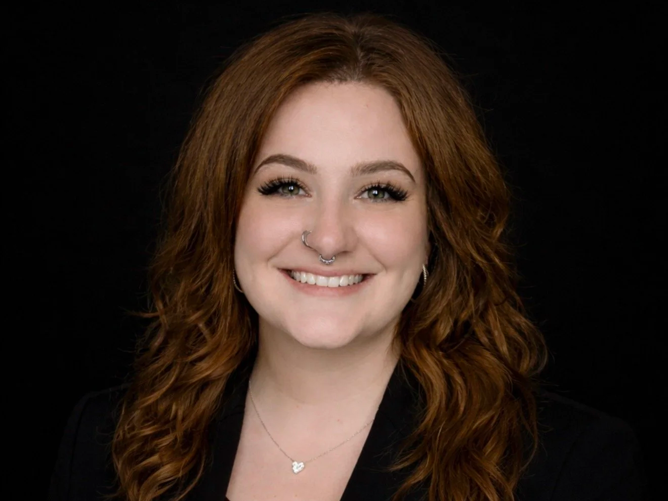 Portrait of a young woman with red hair, nose ring, and a necklace, smiling against a black background.