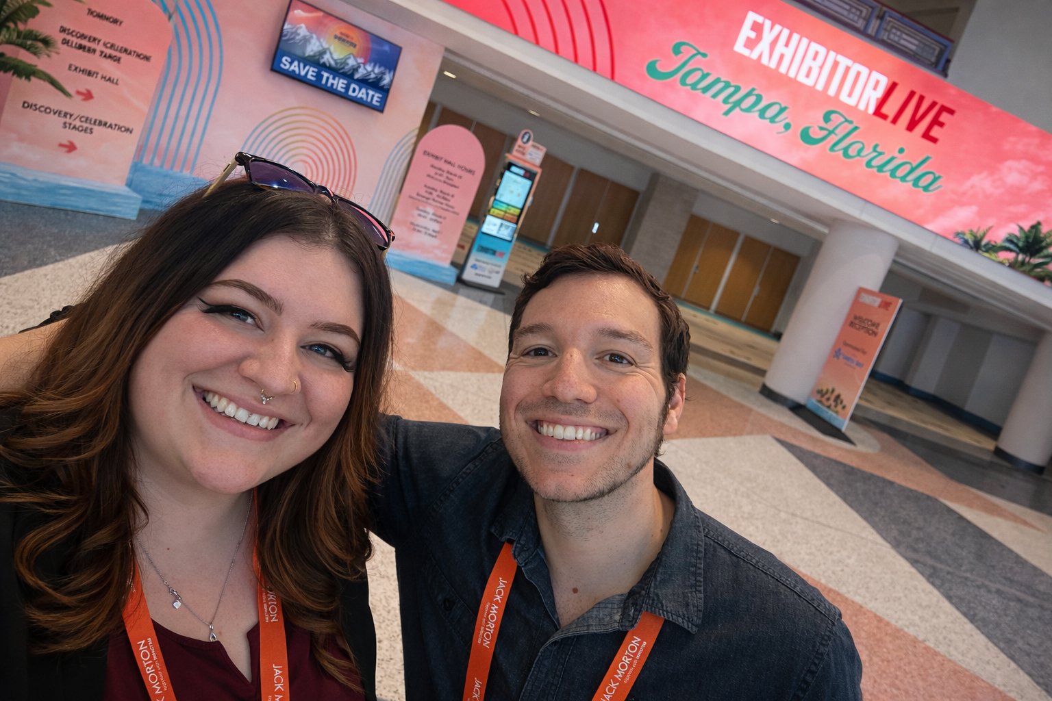A smiling woman with long brown hair, sunglasses on her head, and a nose piercing, taking a selfie with a smiling man with short brown hair at the Tampa, Florida Expo Hall entrance.