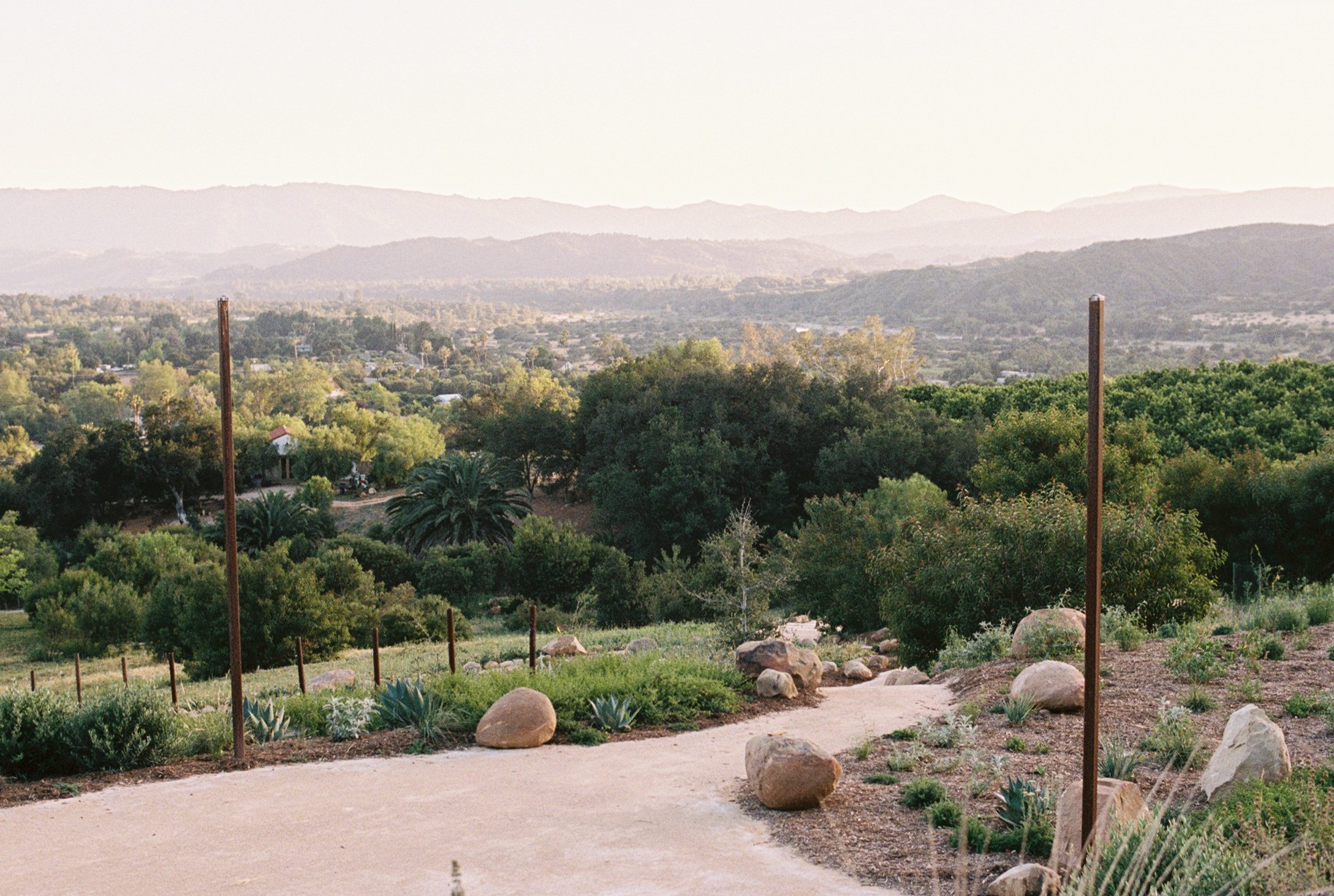 A winding granite path in a lush green landscape with large boulders and succulents, leading to a view of a rural area with trees and distant mountains in the background.