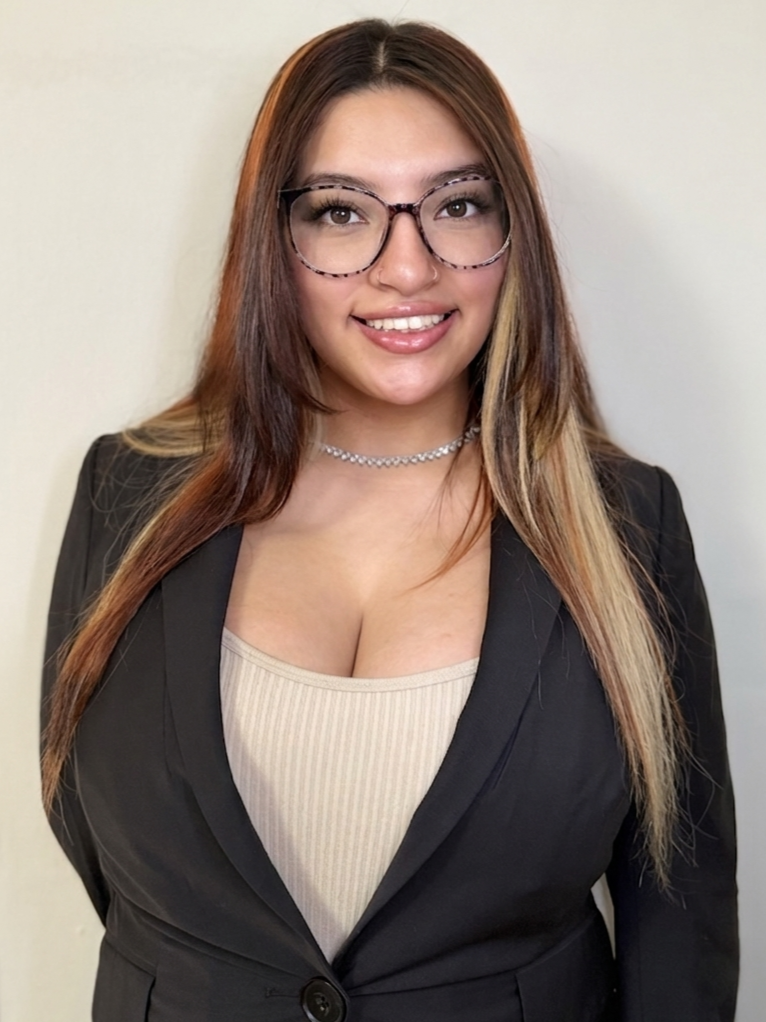 A young woman with long brown hair, wearing glasses, a black blazer, a beige top, a silver choker, and smiling in front of a plain white wall.