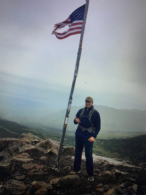 Person standing on rocky terrain holding onto a tall flagpole with an American flag, with a foggy mountainous landscape in the background.