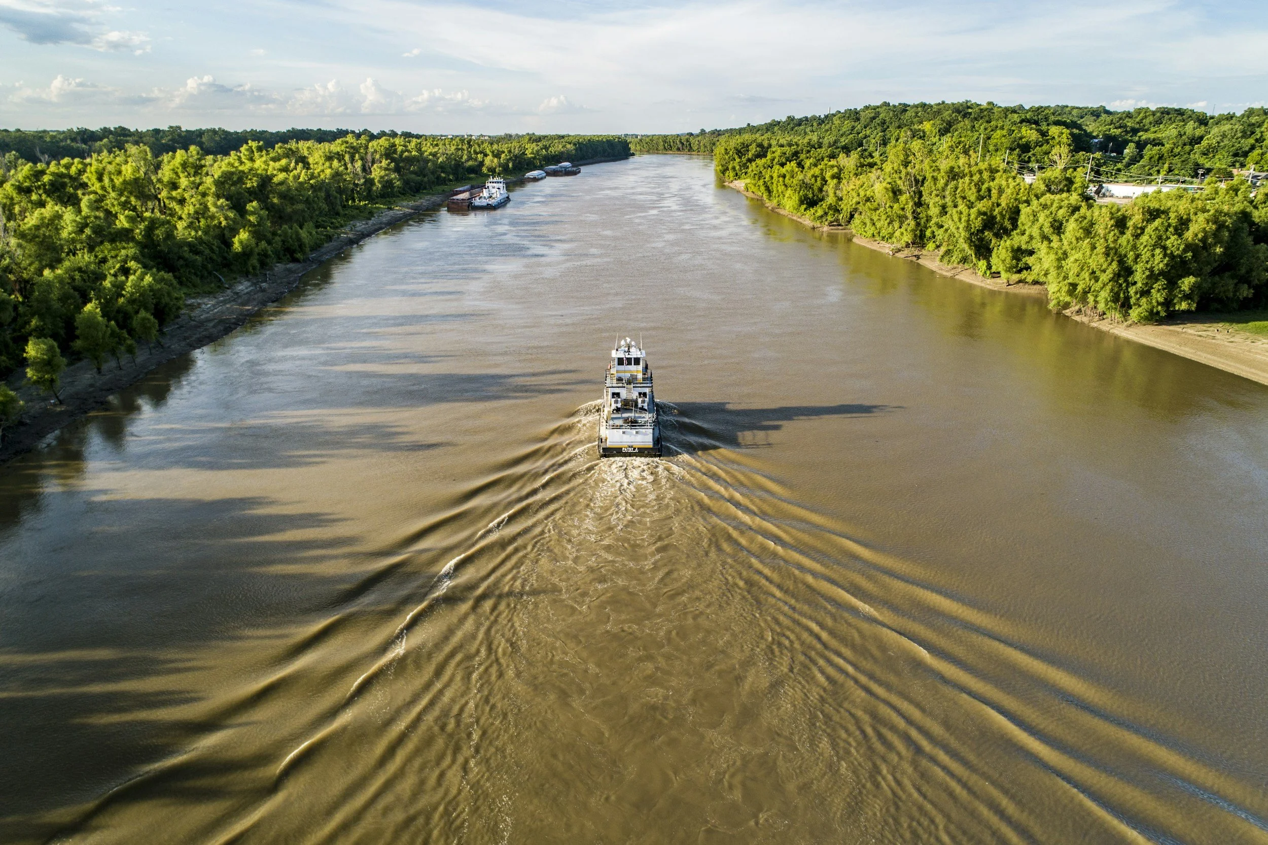 A drone captures a wide view of a river with boats, including a boat in the foreground creating ripples, and lush green trees lining the riverbanks under a partly cloudy sky.