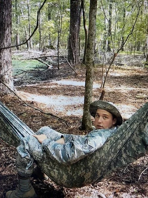 A young person in camouflage military uniform and hat lying in a hammock between trees in a wooded forest.