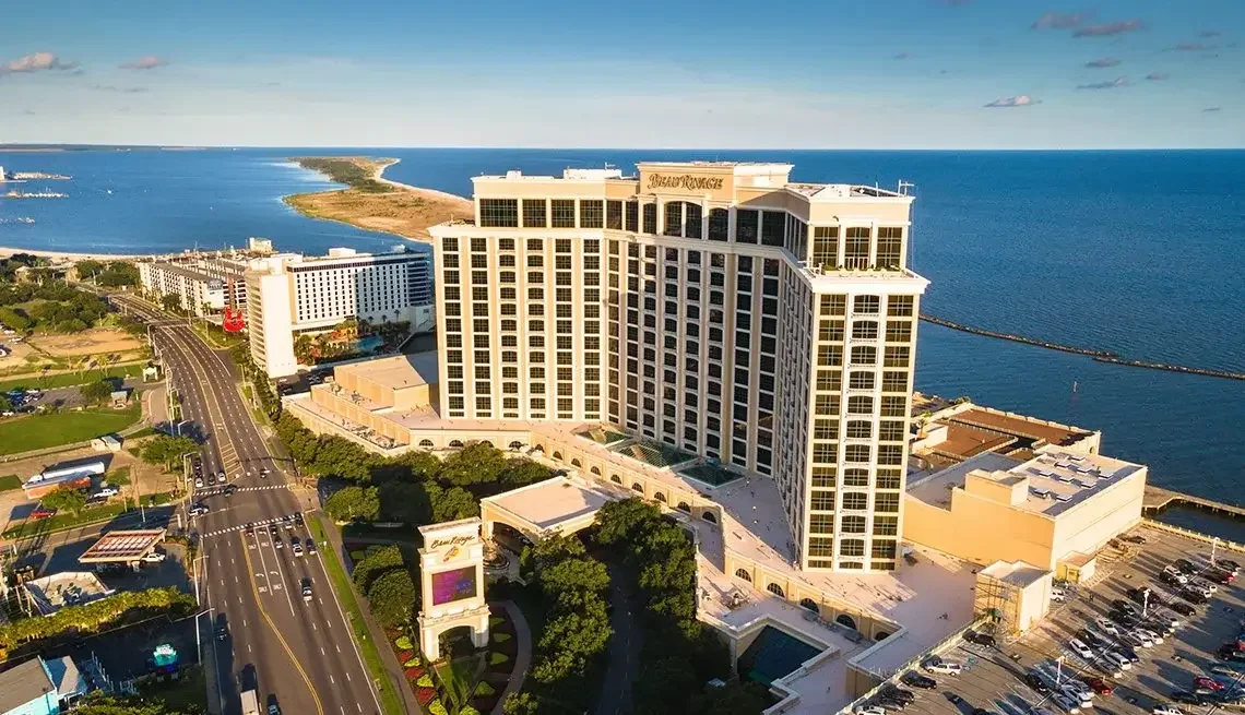 Aerial view of a large hotel by the water with a road and parking lot in the foreground, and a barrier island in the background.