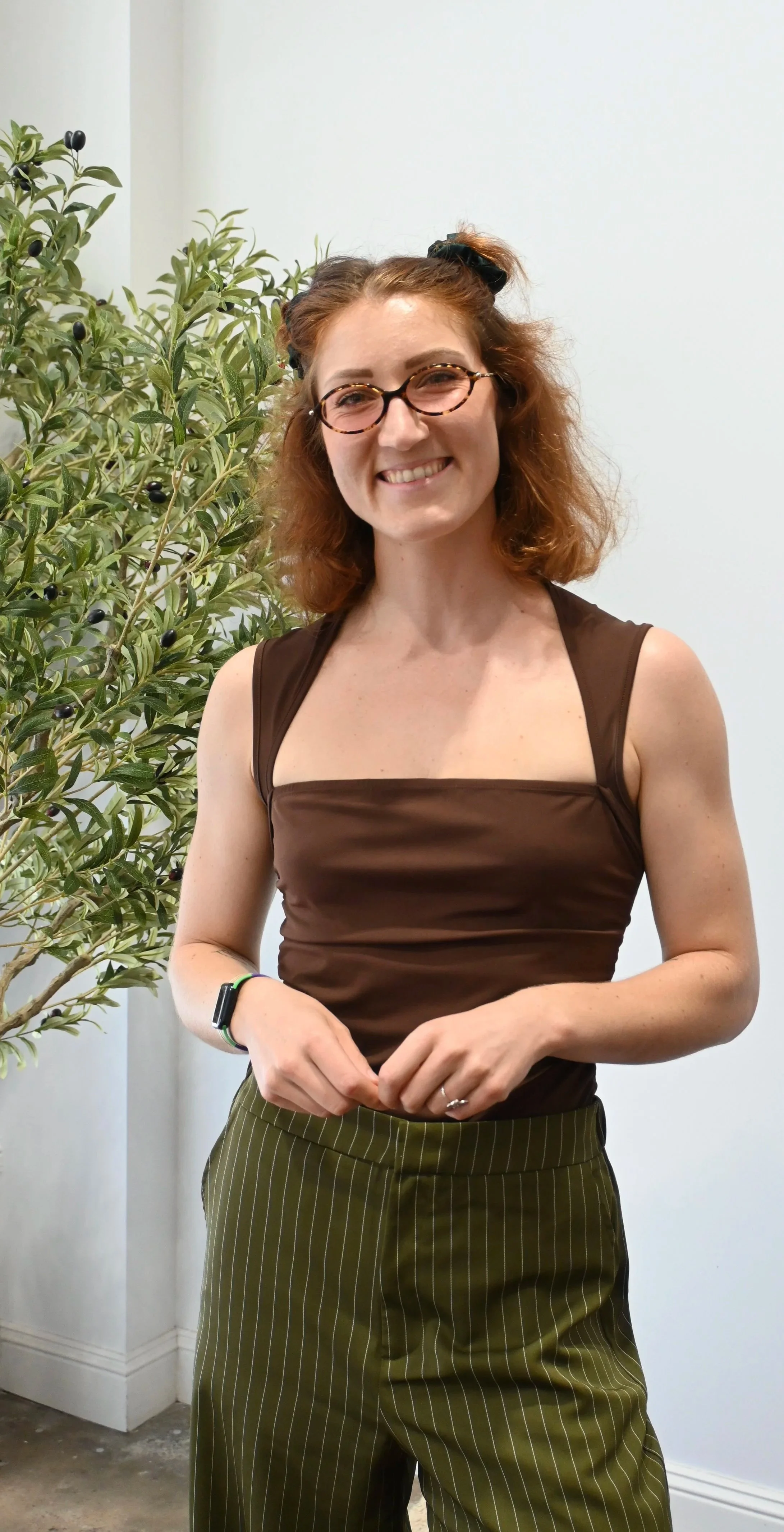A woman with curly red hair, wearing glasses, a brown sleeveless top, and green striped pants, smiling near a green leafy plant with small black berries.
