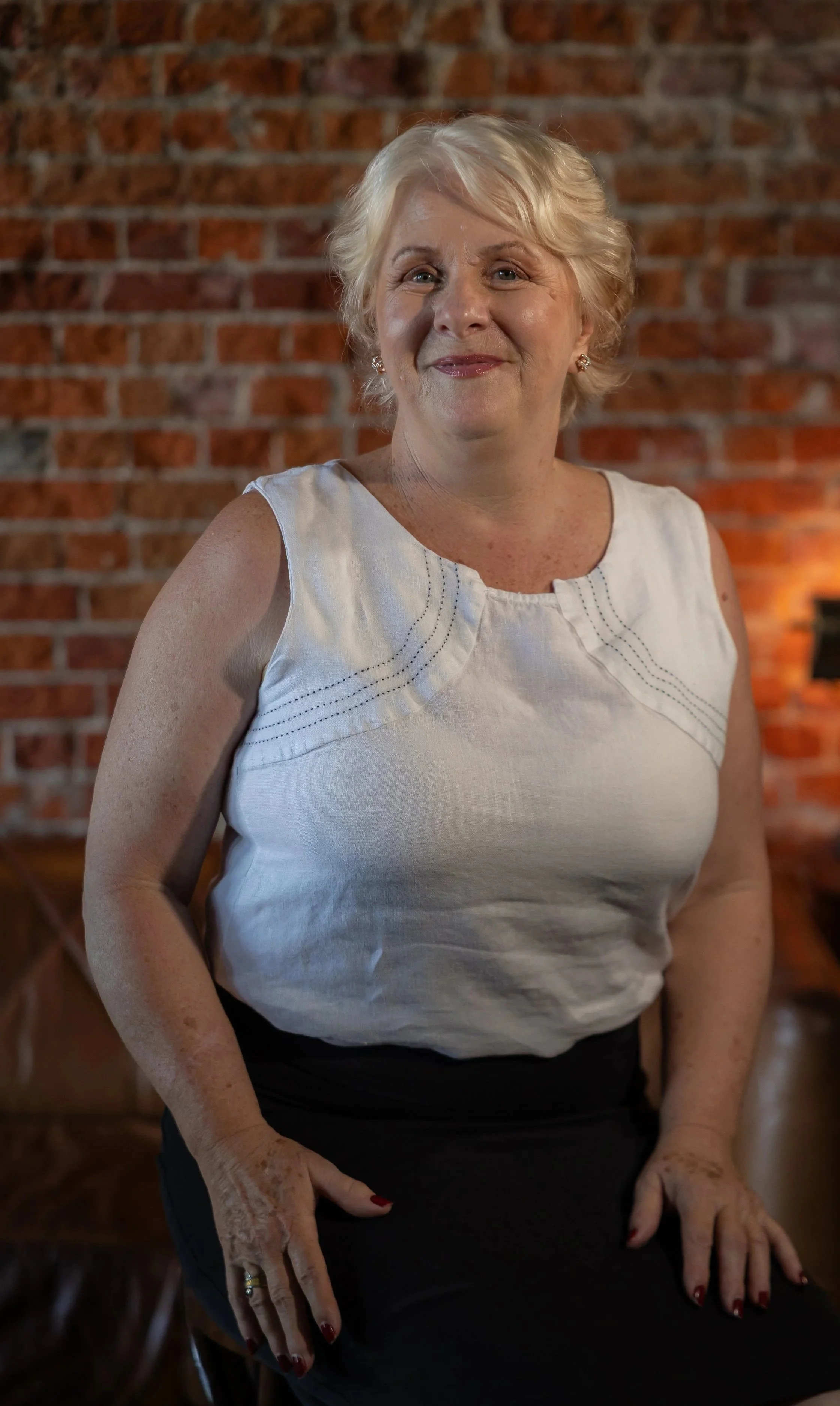 An older woman with short blonde hair, wearing a white sleeveless top and black skirt, sitting against a red brick wall background.