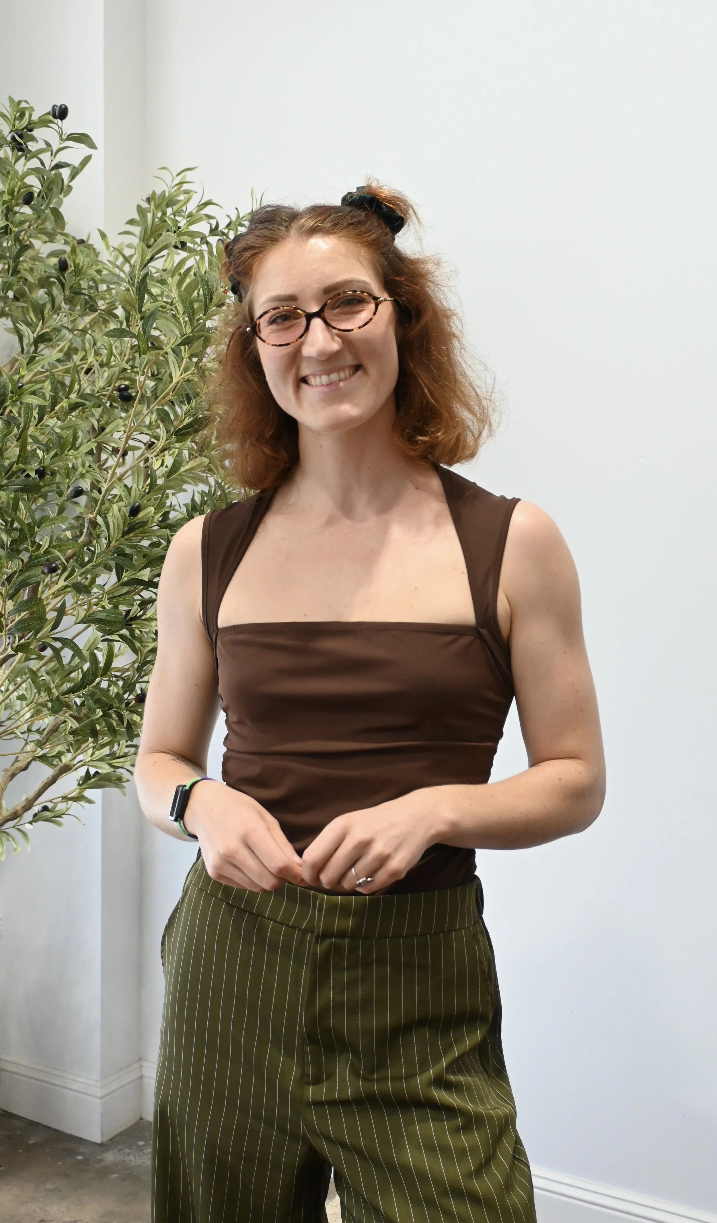 A woman with curly red hair and glasses smiling, wearing a brown sleeveless top and striped green pants, standing indoors next to a green plant with black berries.