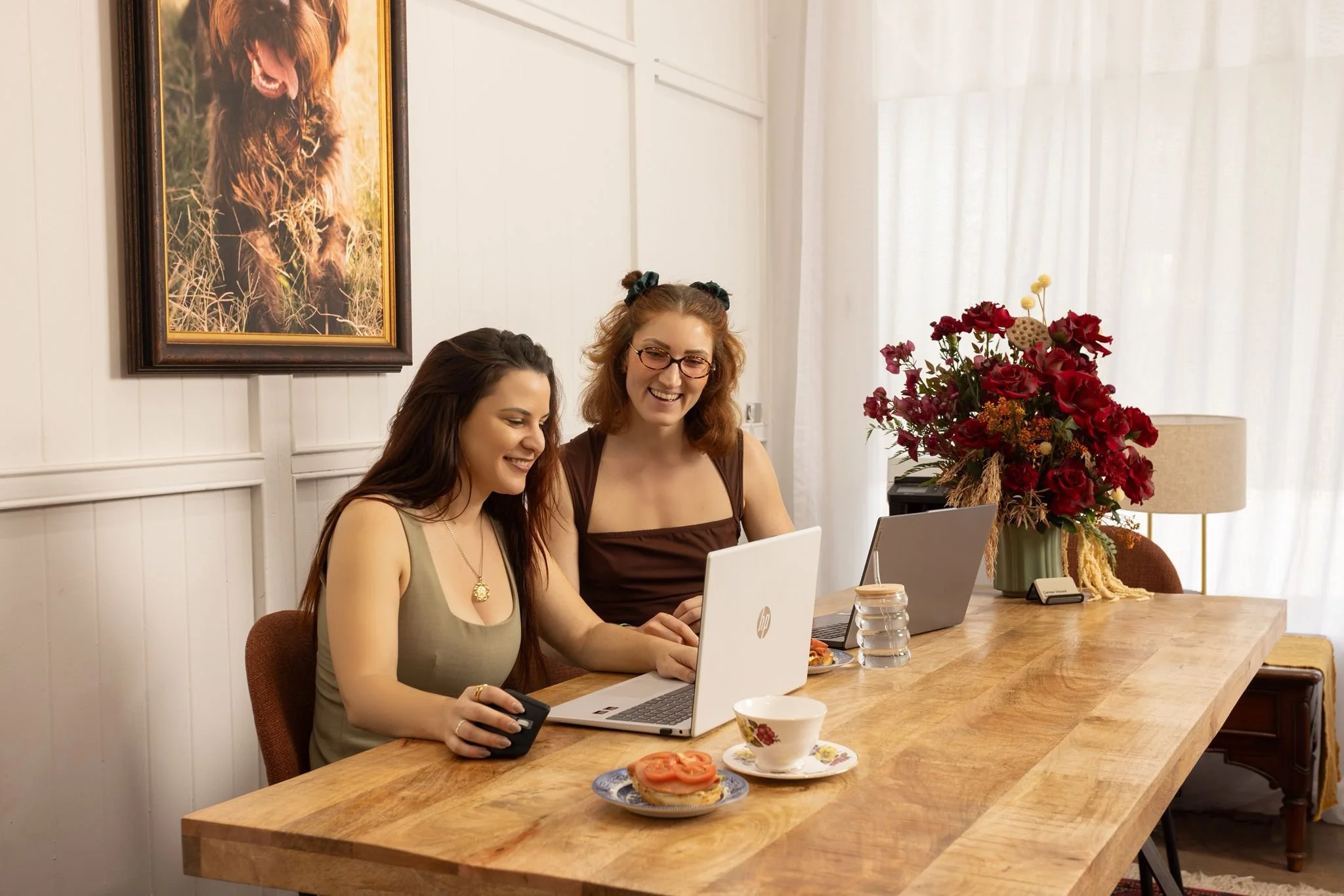 Two women sitting at a wooden dining table, working on laptops with a phone in hand, in a bright room with white paneled walls, a large floral arrangement, and a framed picture of a dog hanging on the wall.