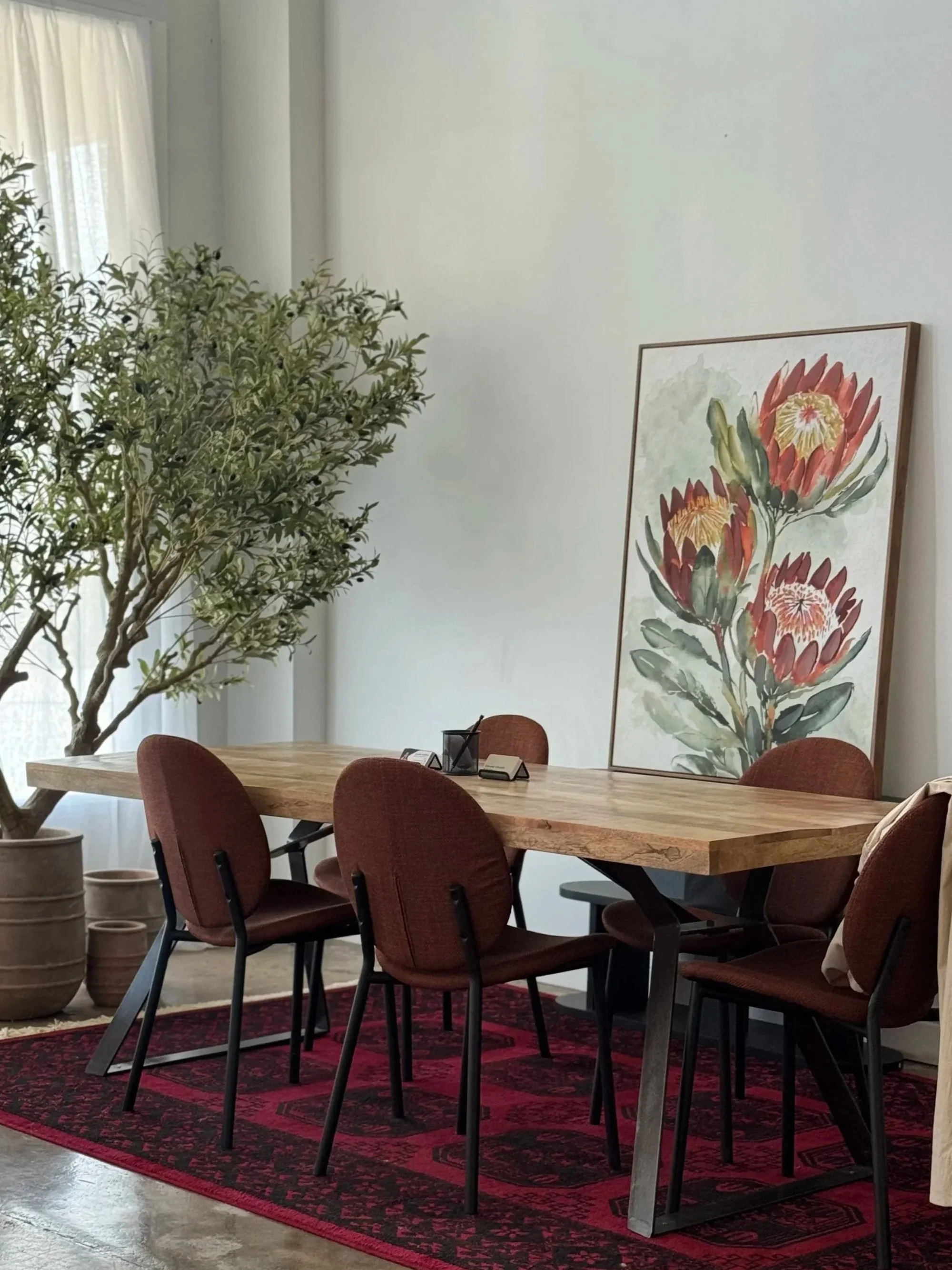 A wooden conference table surrounded by six red upholstered chairs, sitting on a red patterned rug, with a large framed floral painting and a potted plant in the background.