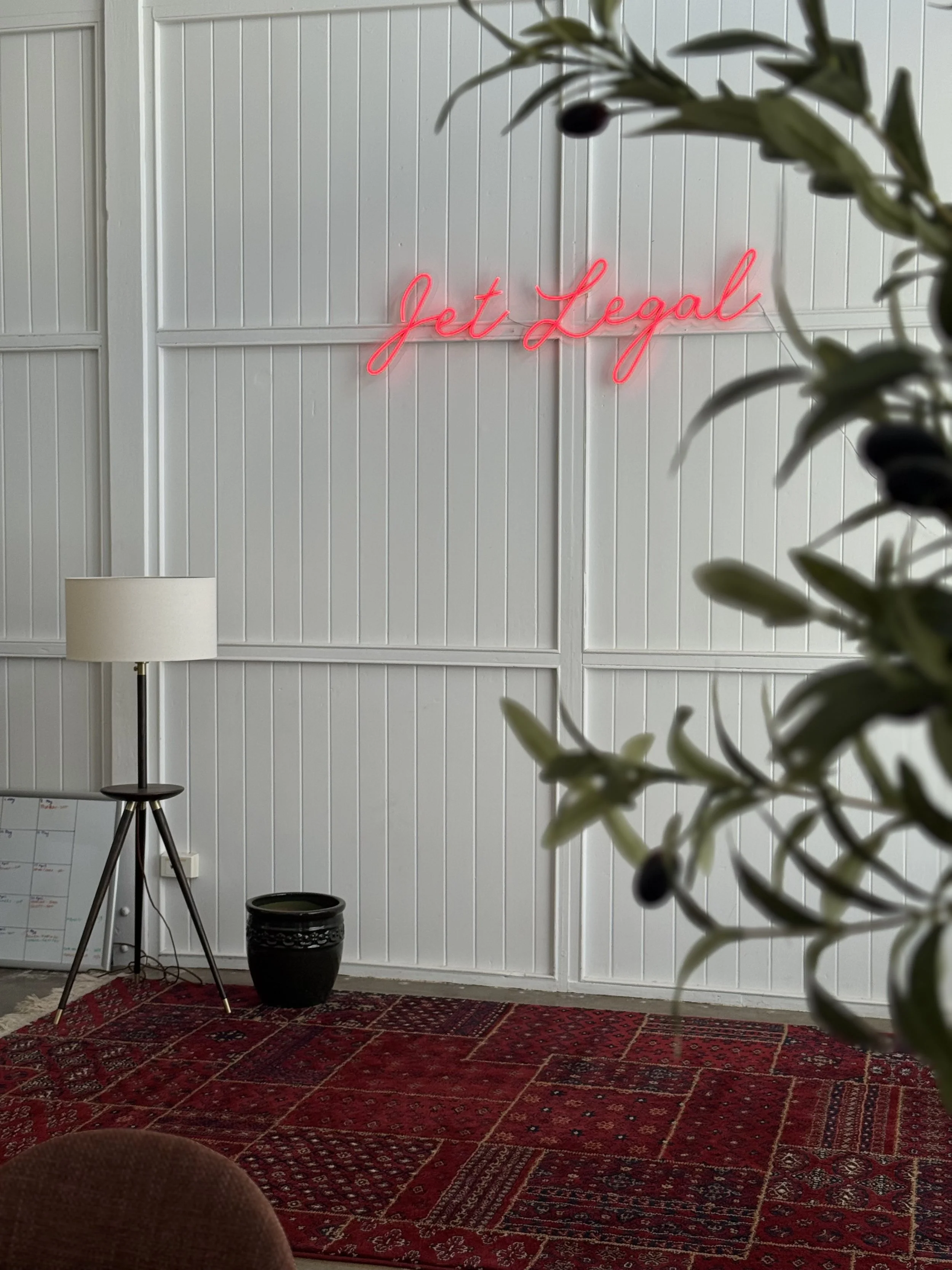 Interior view of a room with white paneled walls, a red patterned area rug, a floor lamp, a potted plant, and a pink neon sign that reads 'Get Legal' on the wall.