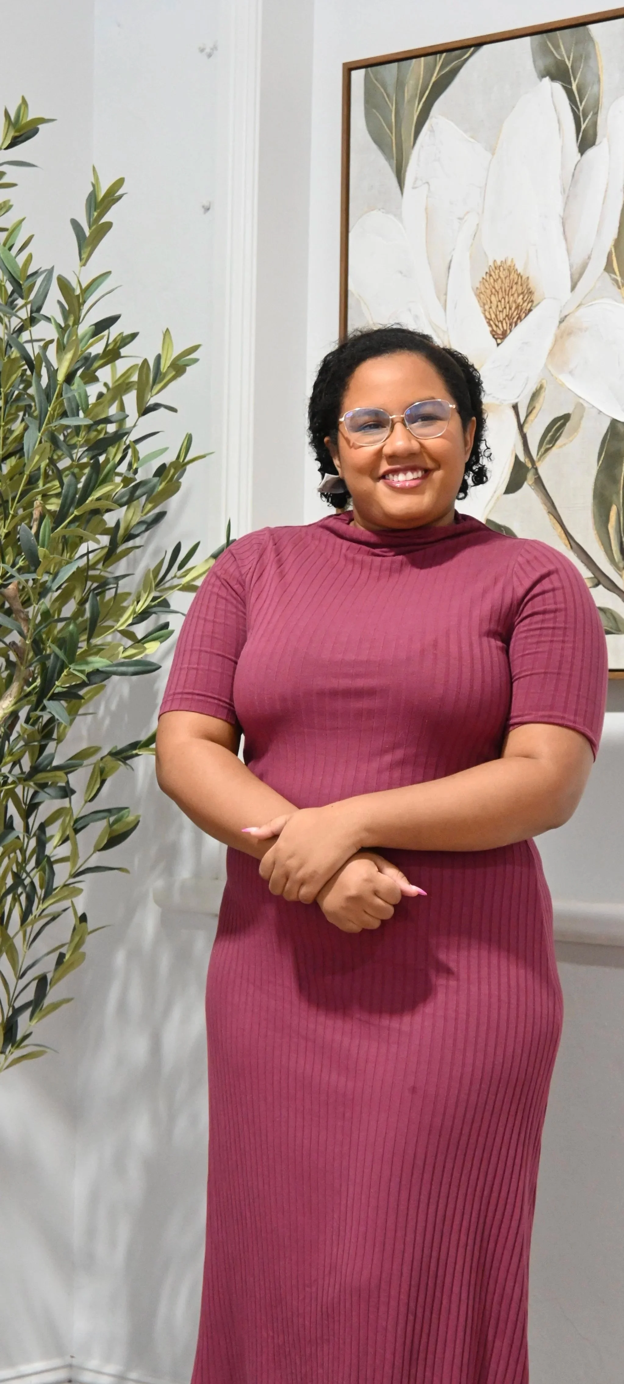 A smiling woman with glasses wearing a maroon ribbed dress standing indoors next to a potted plant and a large framed botanical artwork on the wall.