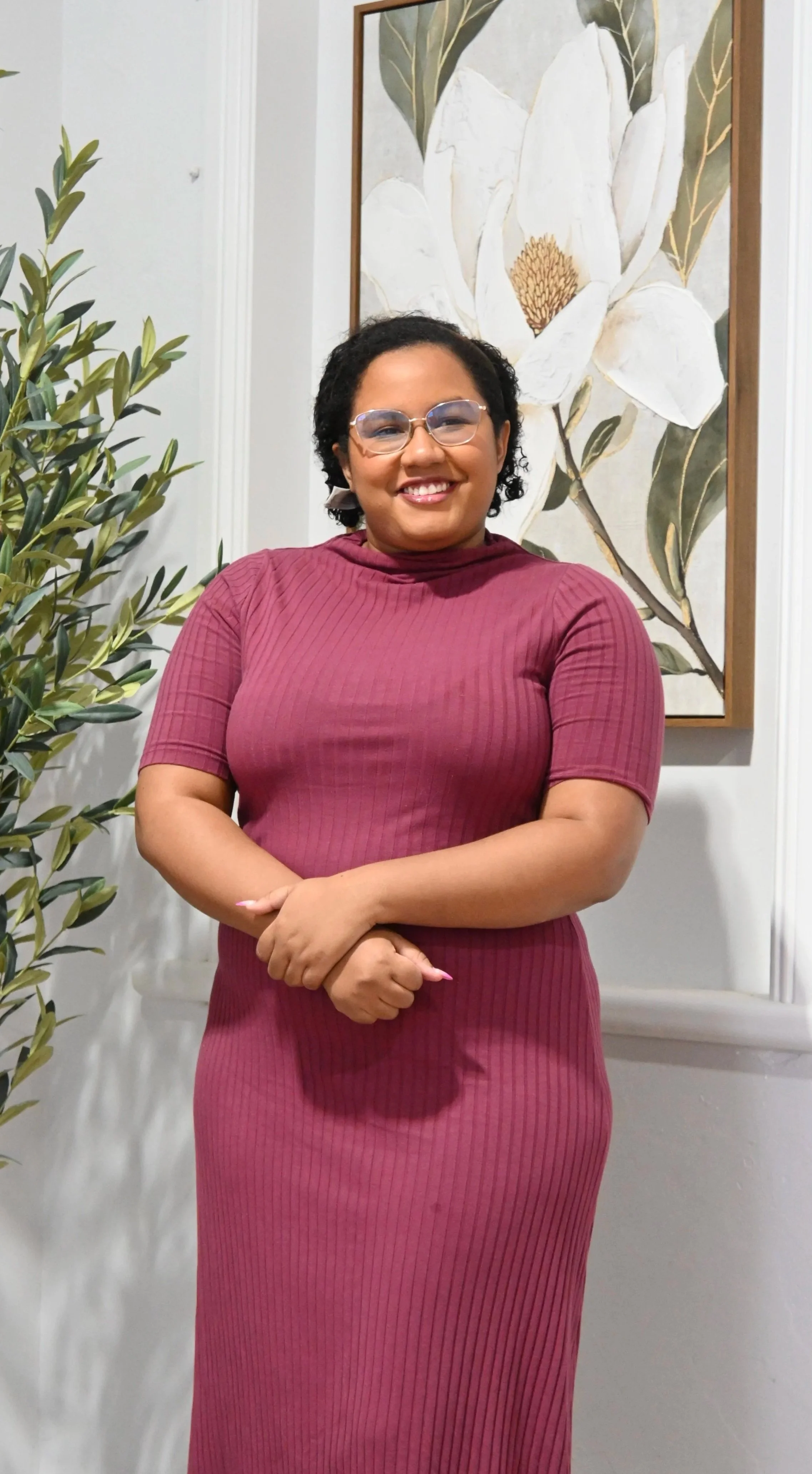 A smiling woman with short curly hair and glasses, wearing a maroon, ribbed, short-sleeved dress, standing indoors next to a large greenery plant and a wall hanging painting of a white magnolia flower.