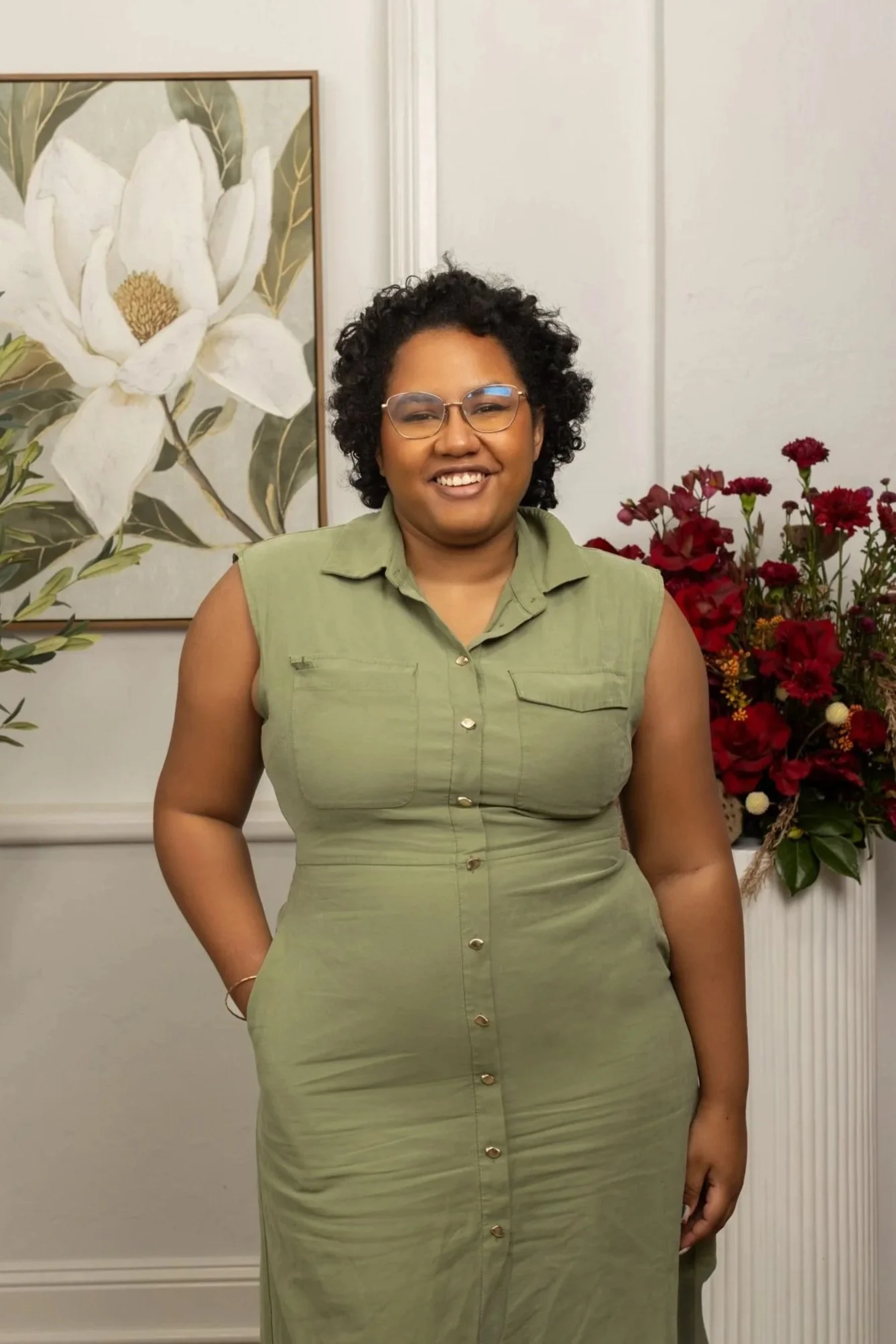 A woman with curly black hair and glasses smiling, wearing a sleeveless olive green button-up dress, standing in front of a wall with floral artwork and a large bouquet of red flowers.