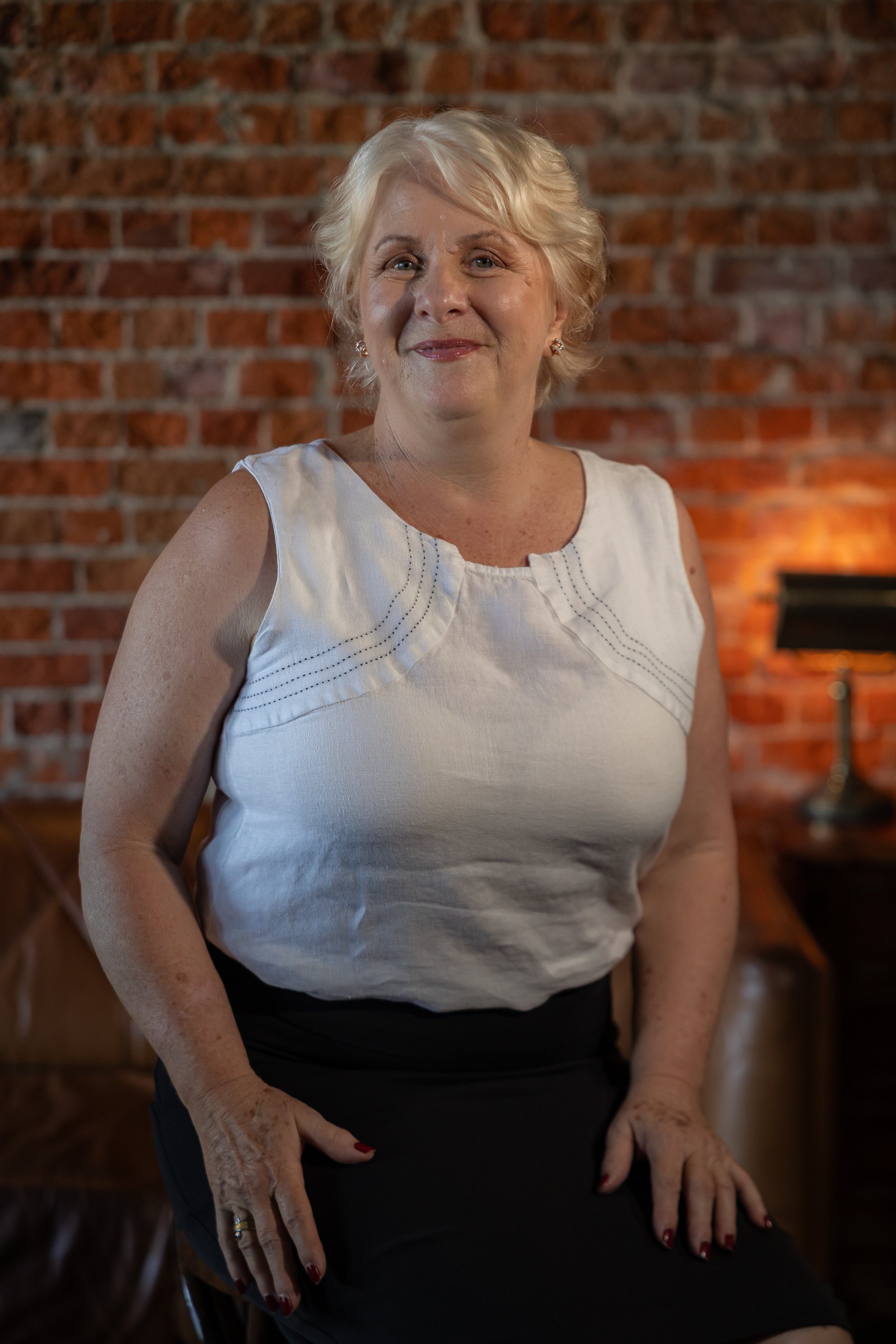 A smiling middle-aged woman with short blonde hair, wearing a sleeveless white blouse, black skirt, and pearl earrings, sitting in front of a brick wall in a warmly lit room.
