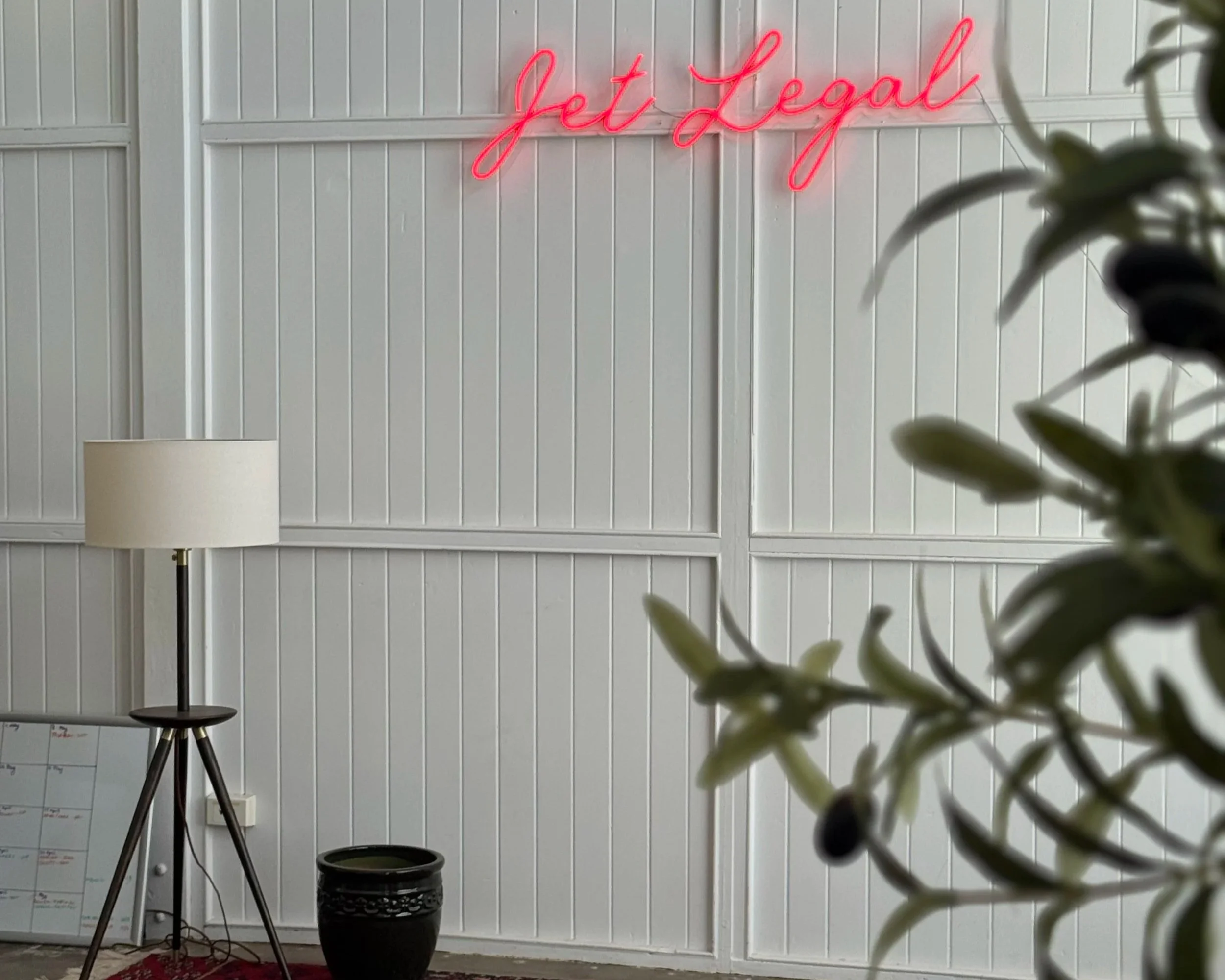 A neon pink sign reading 'Get Legal' hangs on a white wall with vertical paneling. There is a white floor lamp with a beige lampshade to the left, a small potted plant in a dark pot in the center, and some blurry leaves in the foreground on the right.