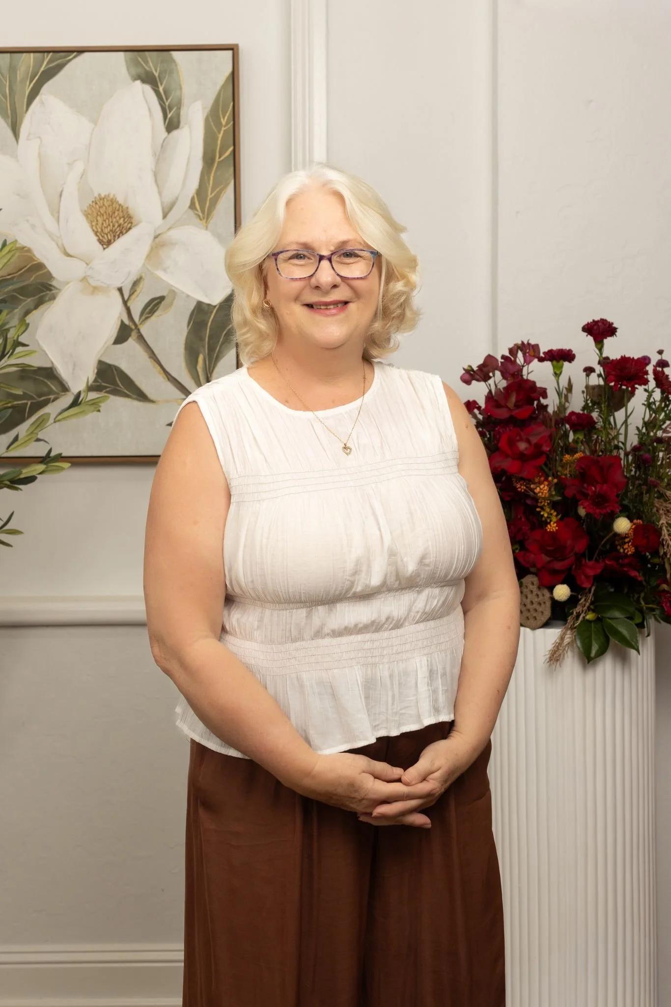 A smiling woman with blonde hair and glasses standing in front of botanical artwork and a floral arrangement.