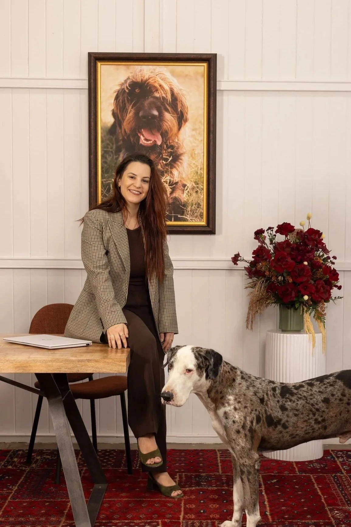 A woman with long brown hair wearing a checkered blazer and dark pants standing beside a large Dalmatian dog inside a room with white paneled walls. Behind her, a large framed photo of a fluffy black and brown dog is hung on the wall. There is a wooden table with a closed laptop on it, a brown chair, a tall white vase with a red and purple flower arrangement, and a red patterned rug on the floor.