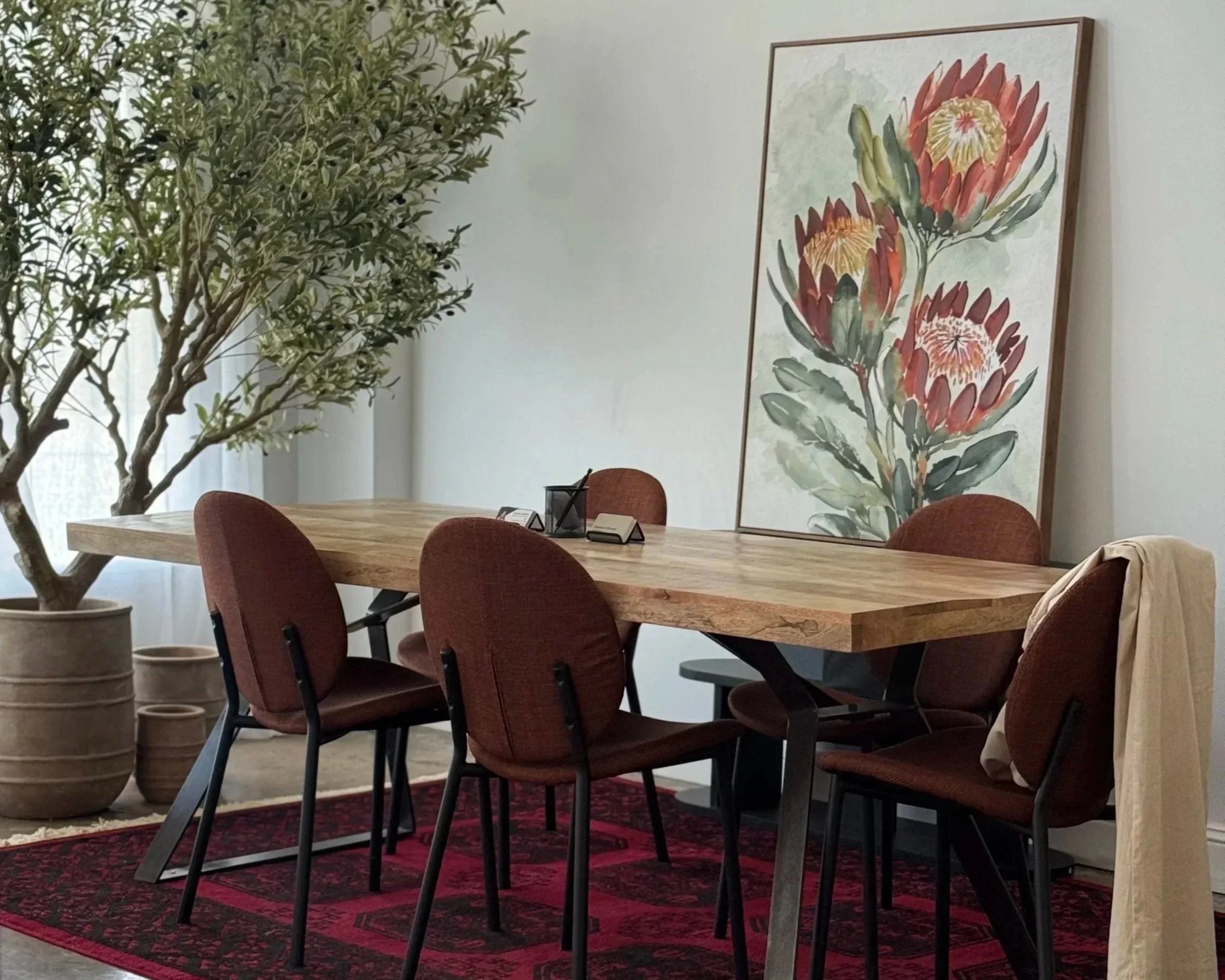 A wooden conference table surrounded by brown upholstered chairs in a room with white walls. A large watercolor painting of orange and yellow flowers decorates the wall. A potted plant with green leaves and small black berries is on the left, and several small desk signs and a lantern are on the table. The floor has a red and black patterned rug.