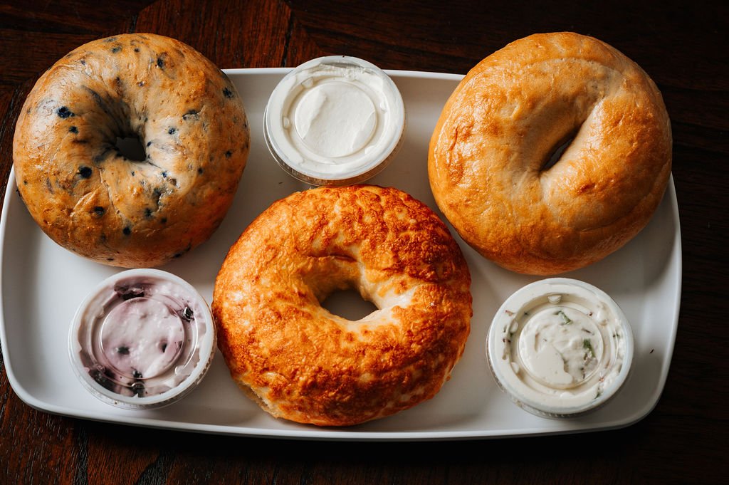 Three bagels with various toppings served with three small containers of flavored house-made cream cheese on a white tray.