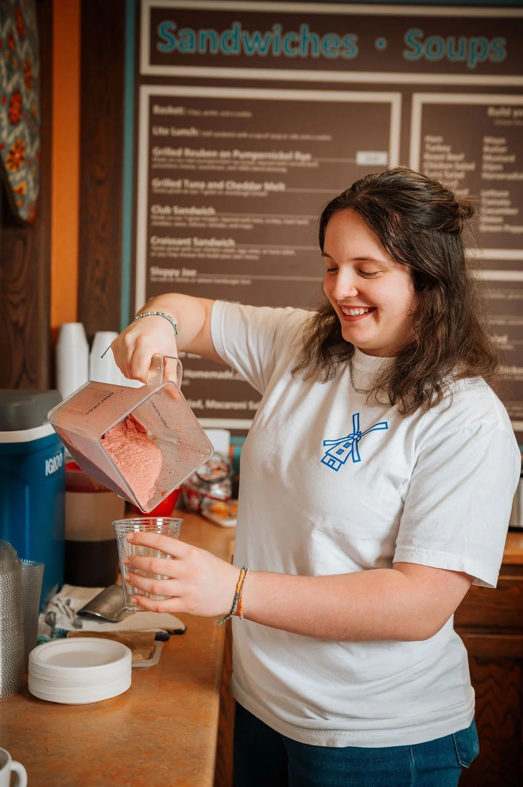 A smiling barista pouring a fruit smoothie into a clear glass at a cafe counter.