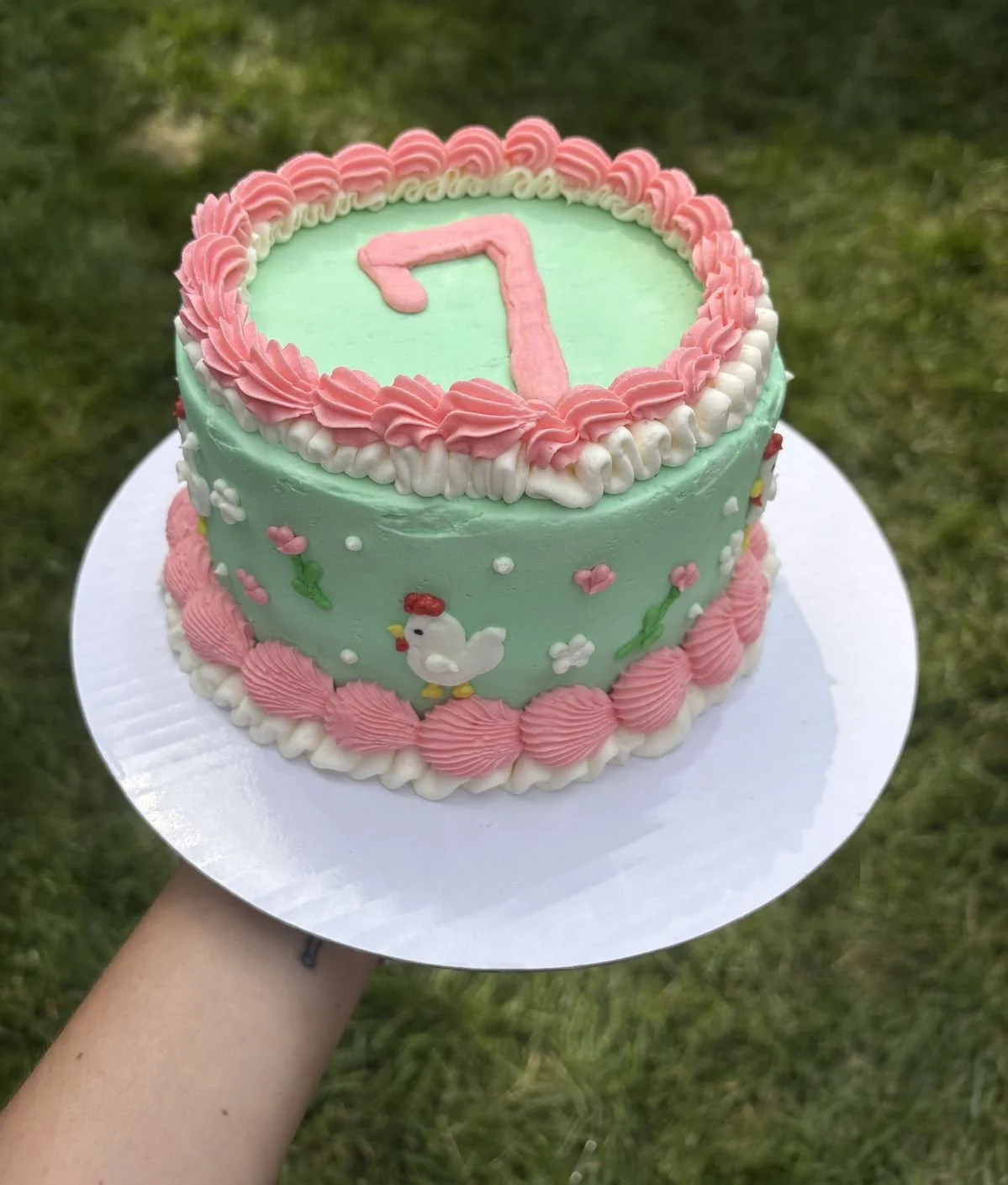 A pastel green birthday cake decorated with pink and white icing, featuring a pink number 7 on top, small flower decorations, and pink icing border accents.