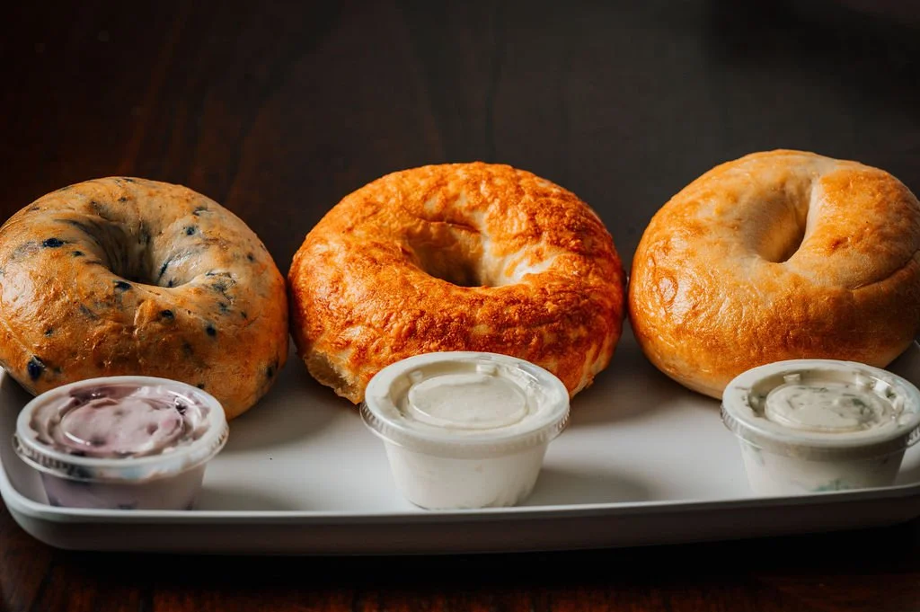 Three bagels with different toppings on a white tray, accompanied by three small containers of house-made cream cheese.