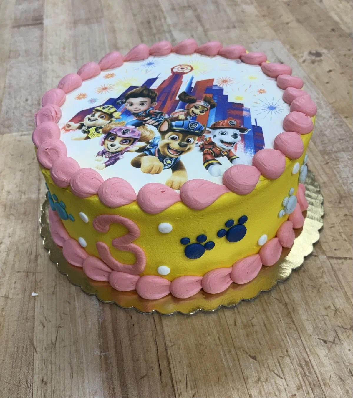 A children's birthday cake decorated with characters from a children's cartoon, featuring a yellow background, pink frosting border, and a printed edible image on top.