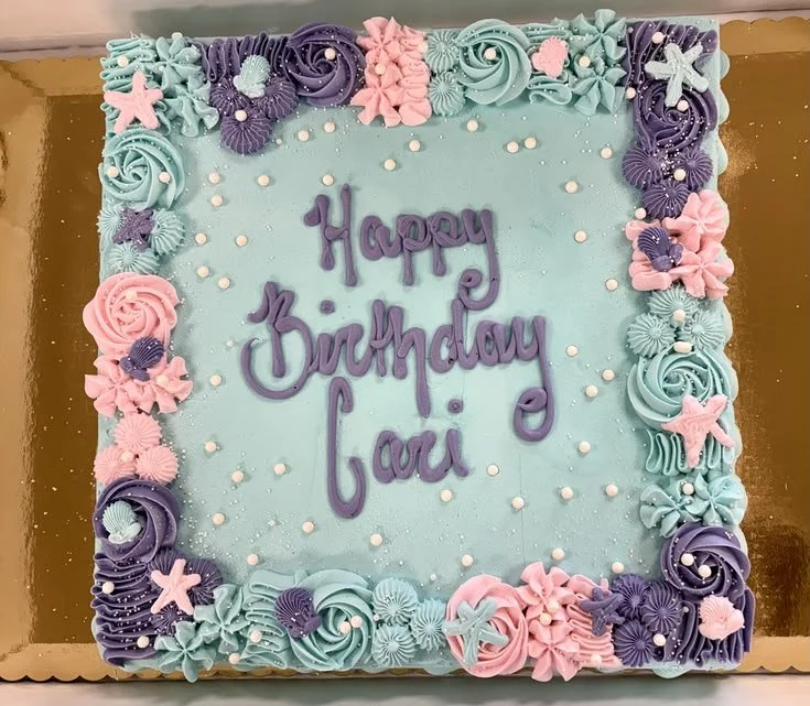 Square birthday cake with pastel blue frosting, decorated with pink, purple, and mint green icing flowers and shells along the border. The center reads "Happy Birthday" in purple icing, with small white edible dots scattered on the surface.