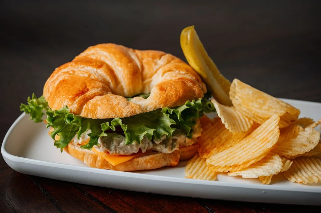 Chicken salad croissant sandwich with lettuce and cheese on a white plate with potato chips and a pickle spear.