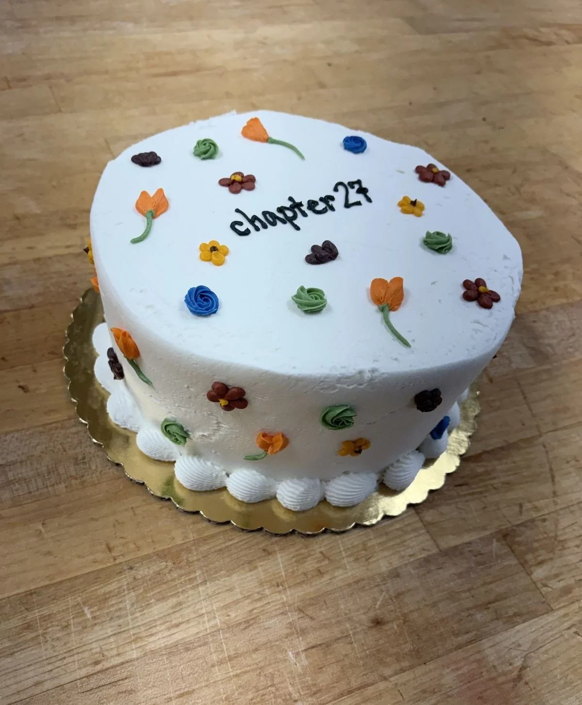 Heart-shaped birthday cake with white frosting, decorated with small colorful flower and leaf-shaped icing, has the text 'chapter 23' written on top, and is placed on a gold cake board on a wooden surface.