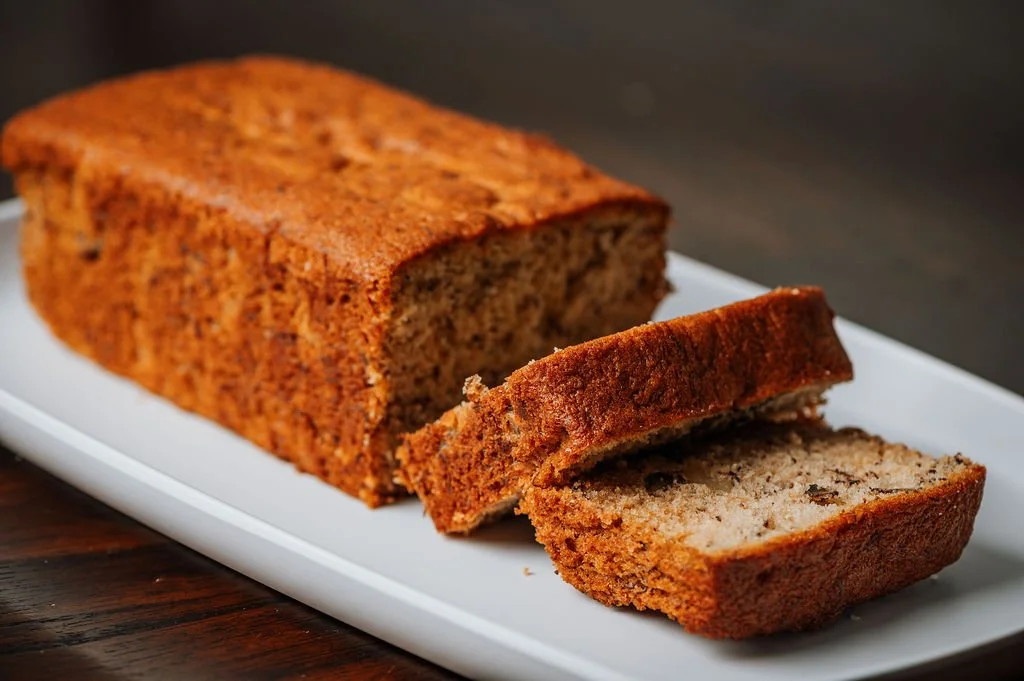 Loaf of banana bread on a white platter with one slice cut and placed in front, showing a moist interior with banana chunks.