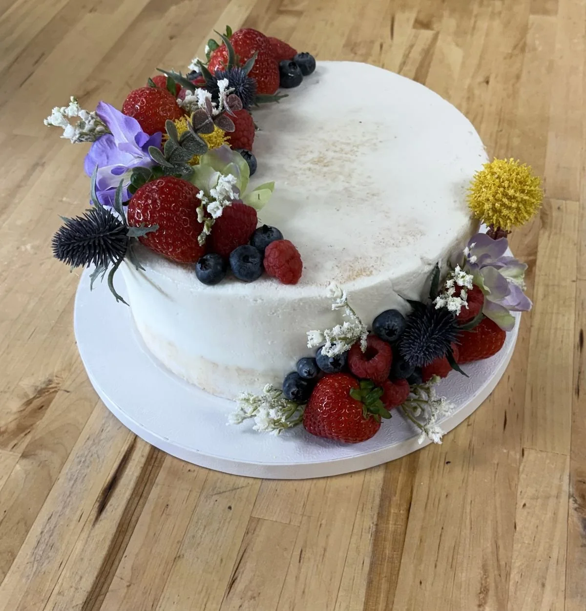 Round white cake decorated with fresh berries and flowers on a white cake board on a wooden table.