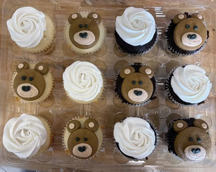 A plastic container holding twelve cupcakes arranged in four rows of three, decorated as bear faces and plain frosting swirls.