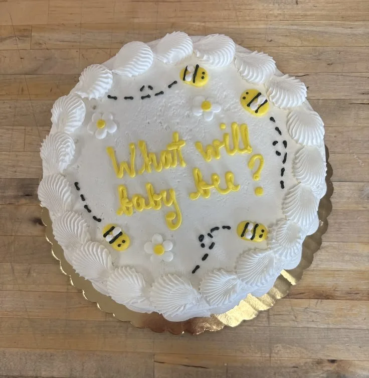 Round white cake with piped white icing border, decorated with small yellow and black bee figures, white flowers with yellow centers, and black dotted lines. Centered yellow writing that reads, 'What will baby be?' on a wooden surface.