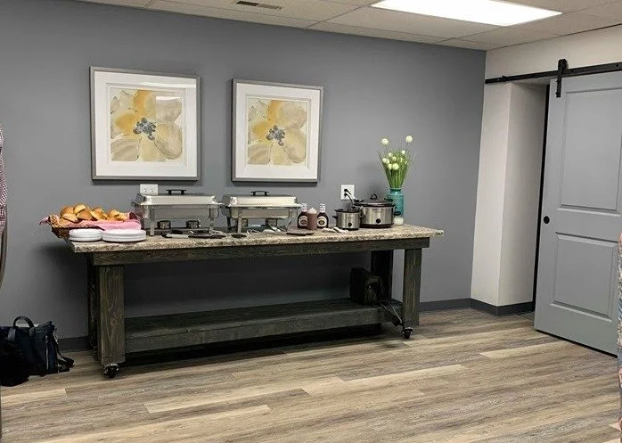 Buffet table with chafing dishes and a tray of muffins in a room with gray walls and wooden flooring.