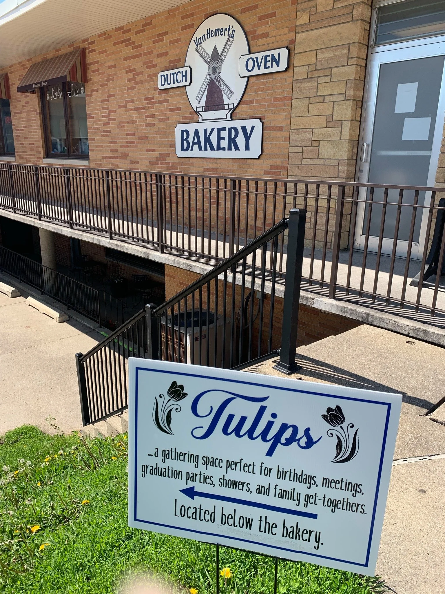 Photo of a bakery sign on a brick wall that reads "Van Hemert's Dutch Oven Bakery" with a windmill illustration. In the foreground, a sign promotes a gathering space for birthdays, meetings, graduation parties, showers, and family get-togethers, loca