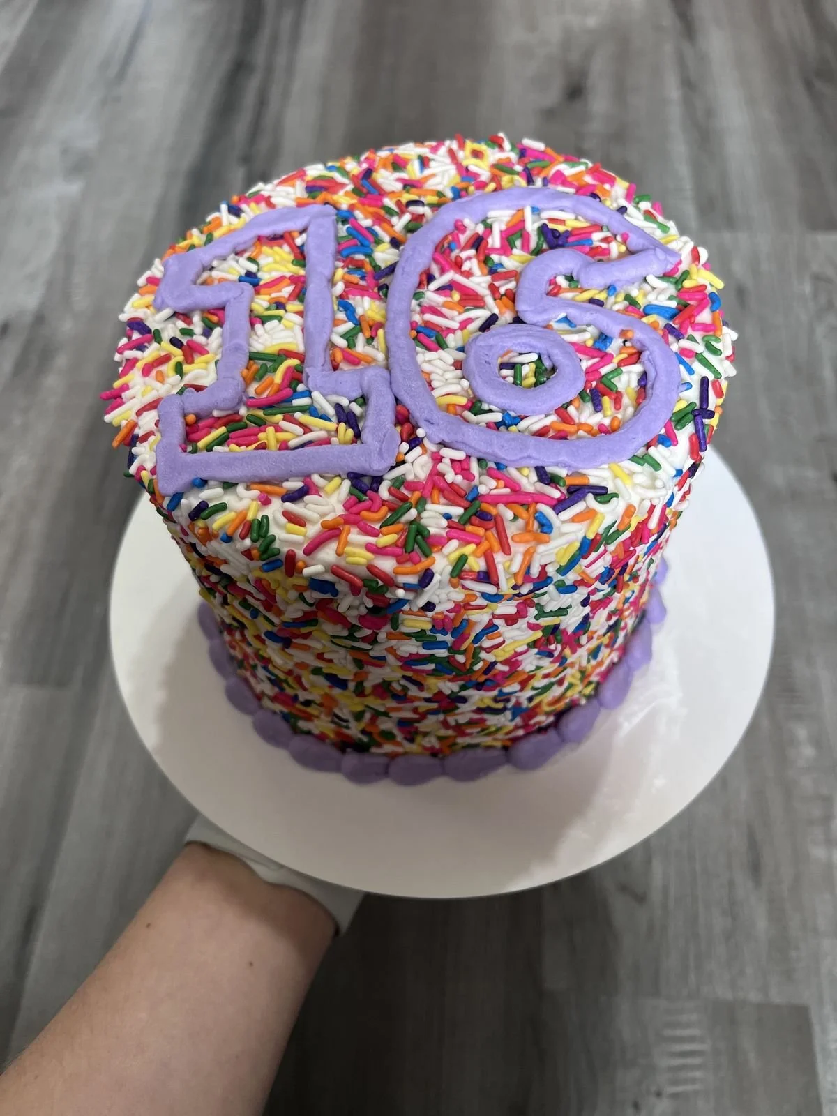 Colorful birthday cake with sprinkles, decorated with purple icing forming the number '20' on top, and it is being held over a white cake stand.