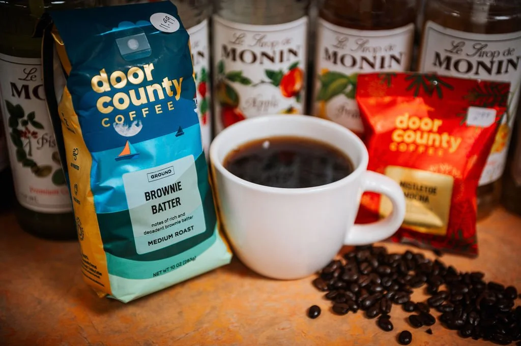 A cup of black coffee surrounded by bags of Door County Coffee and Monin syrup bottles, with scattered coffee beans on a wooden surface.