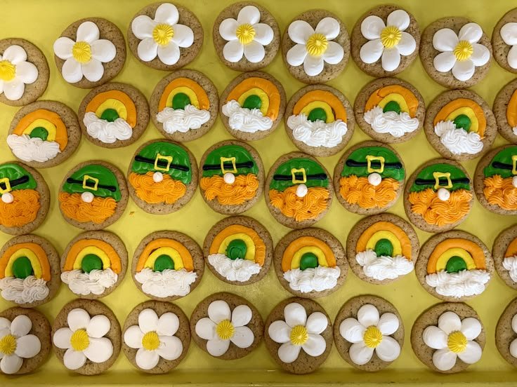 Decorated cookies with three different designs arranged on a yellow surface: white daisy-shaped cookies with yellow centers, rainbow cookies with clouds, and leprechaun hats with orange beards and green hats.