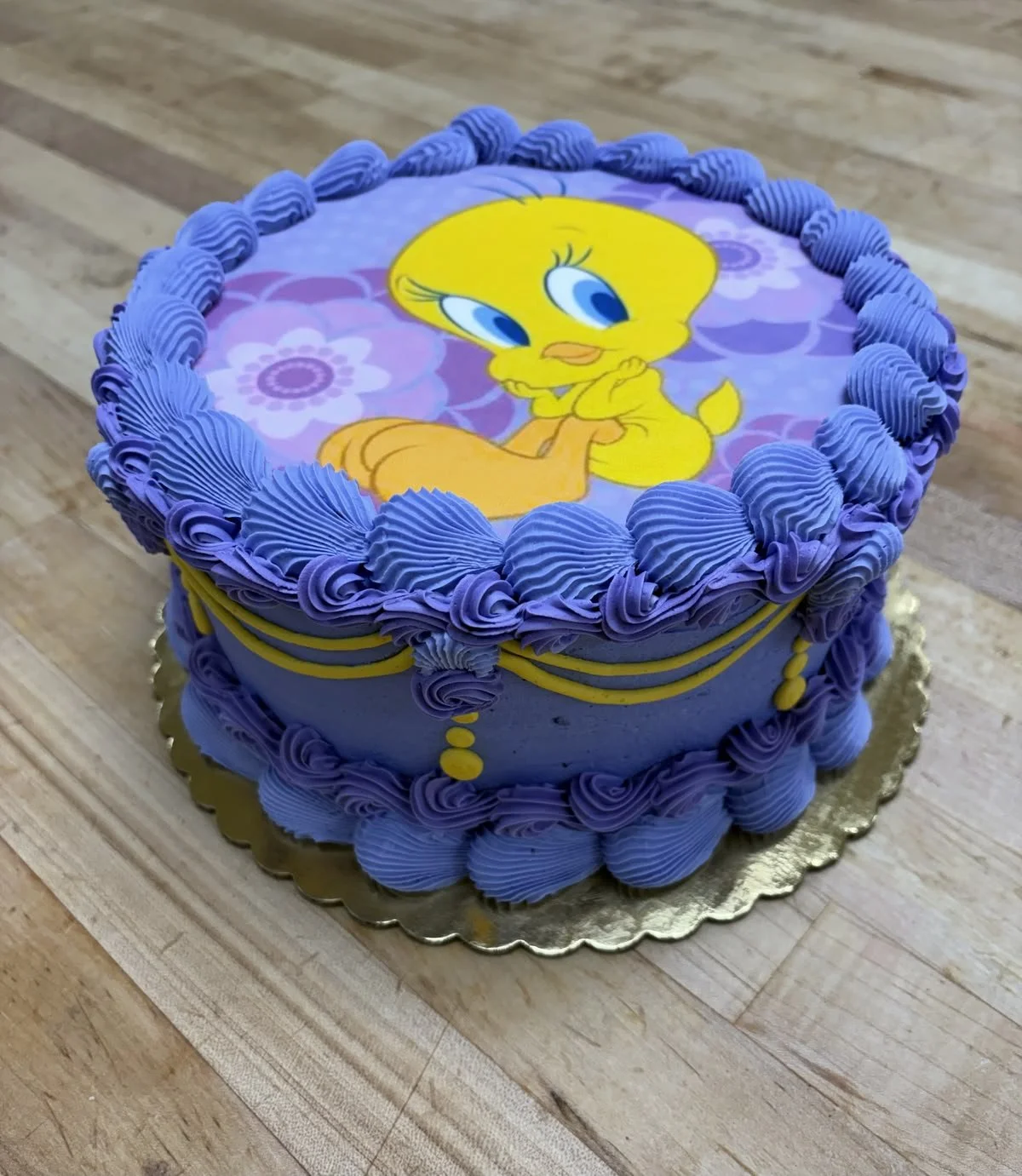 A round cake decorated with purple and yellow frosting, featuring an edible image of Tweety Bird on top, with purple frosting ruffles along the edges.