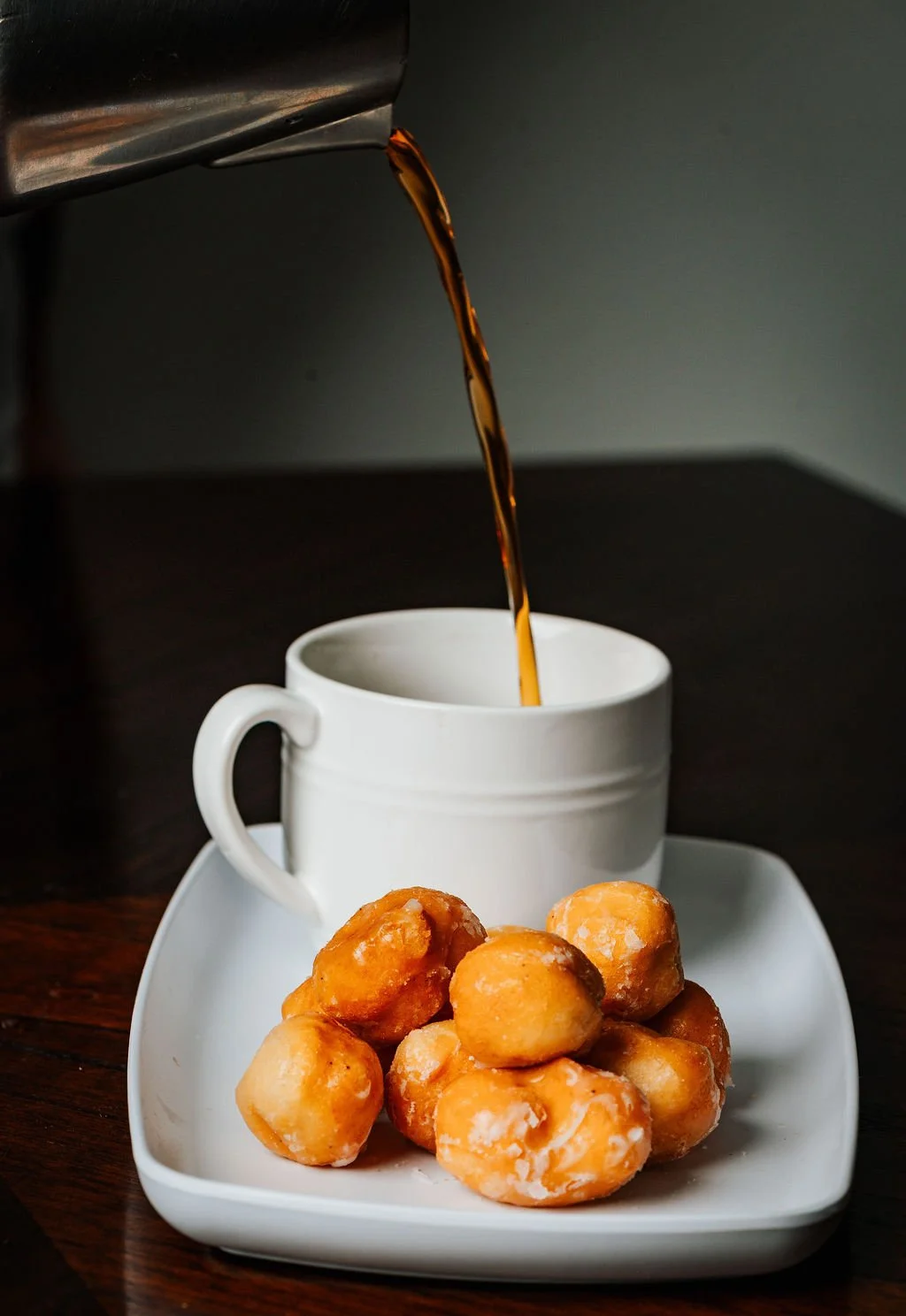 Pouring hot coffee into a white mug with a pile of donut holes on a white plate in the foreground.